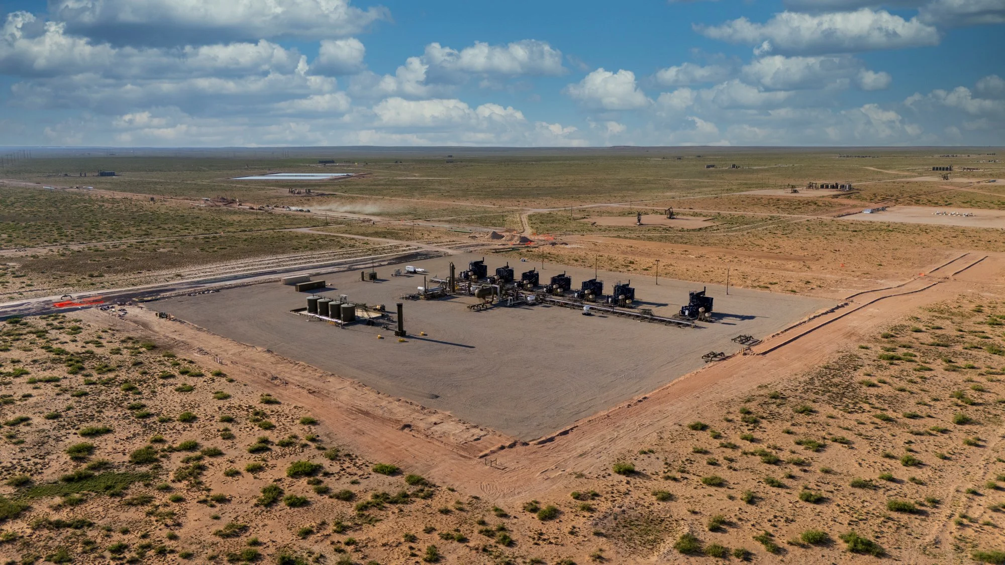Aerial view of an oil or gas drilling site in a desert landscape, with equipment, pipelines, and a few structures under a partly cloudy sky.