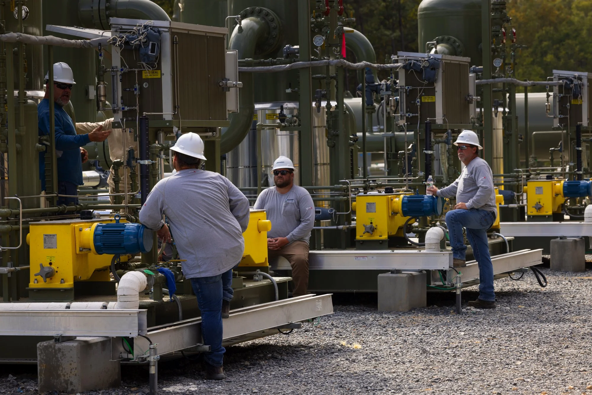 Four workers in hard hats and safety glasses on an industrial site with pipes and machinery, engaging in a discussion or inspection.