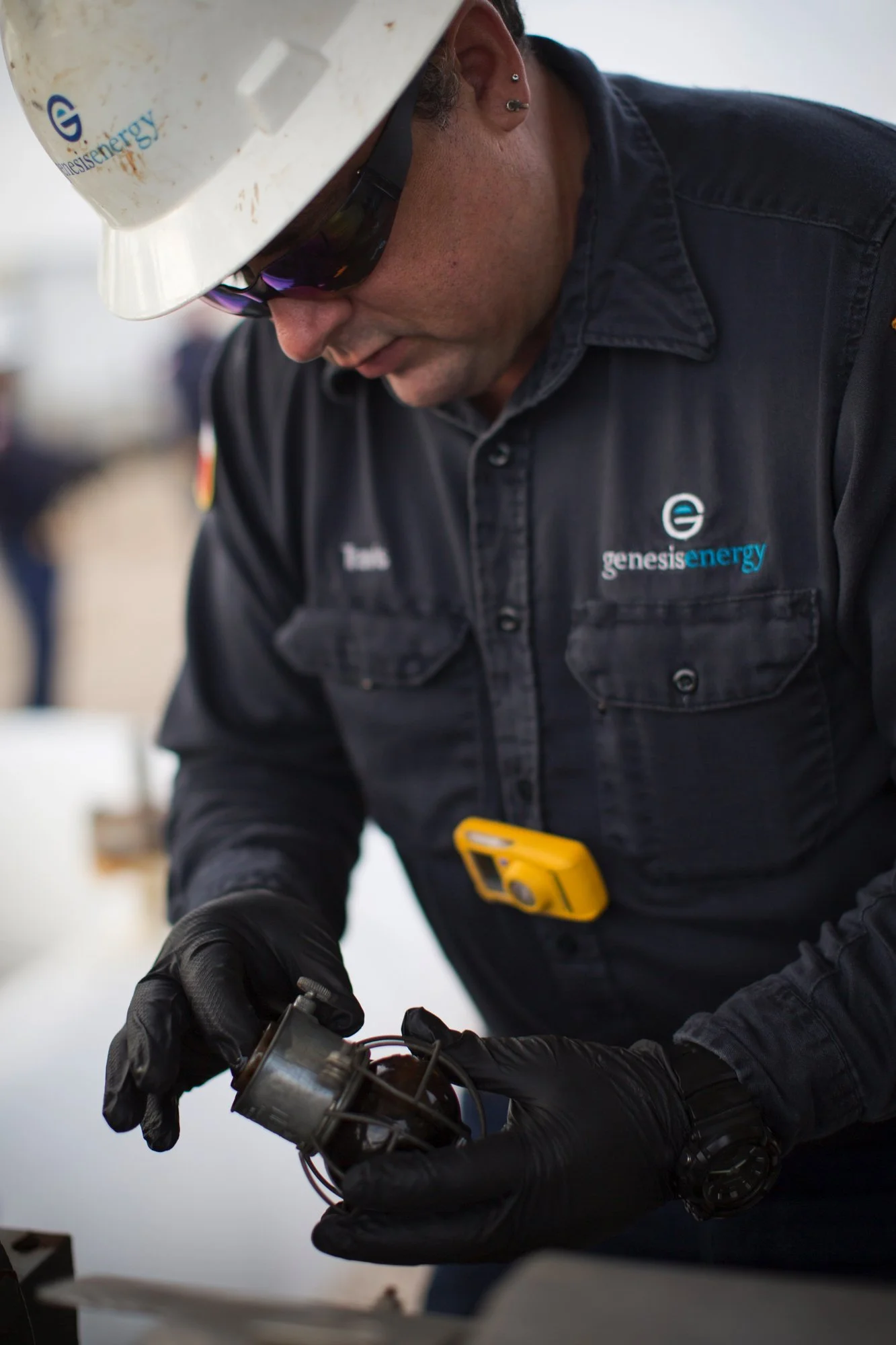 A technician wearing safety glasses, a hard hat with logo, black gloves, and a Genesis Energy shirt inspecting a small mechanical part in a workshop.