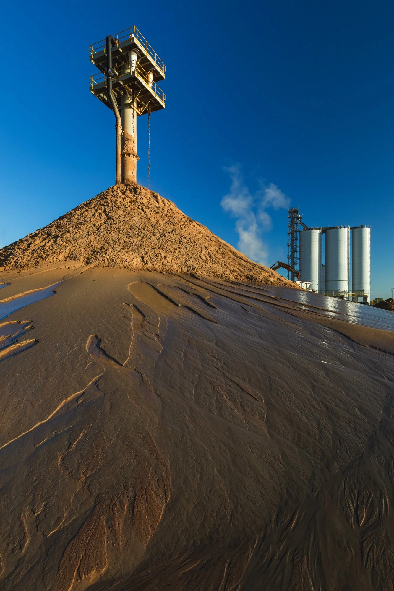 Industrial site with a mound of sand or mud and a tall metal structure on top, with pipes and silos in the background under a clear blue sky.