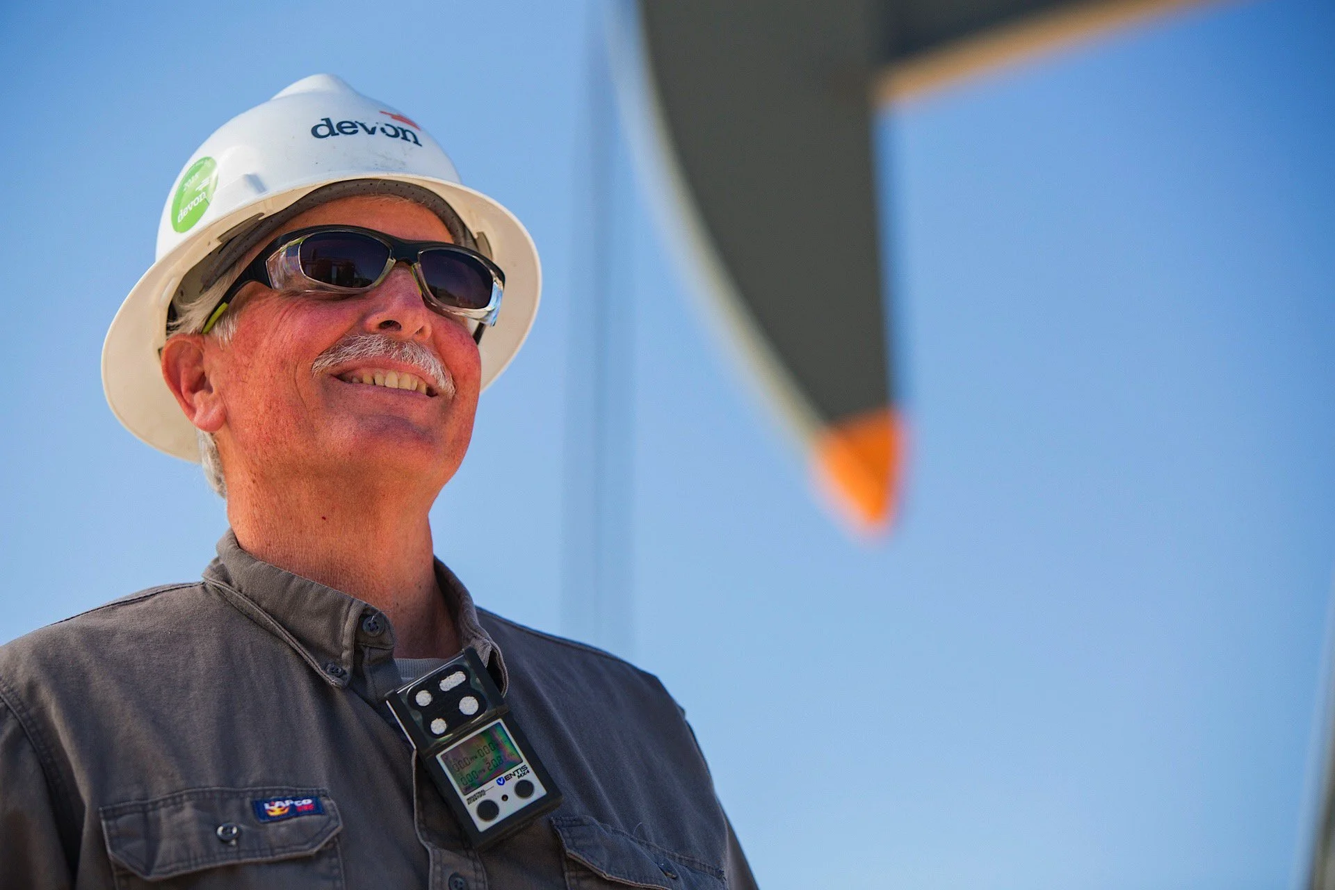 A smiling man wearing sunglasses, a white hard hat with the word 'devon' on it, and a gray work shirt with a worker badge. He is outdoors, possibly at a construction site or industrial area.