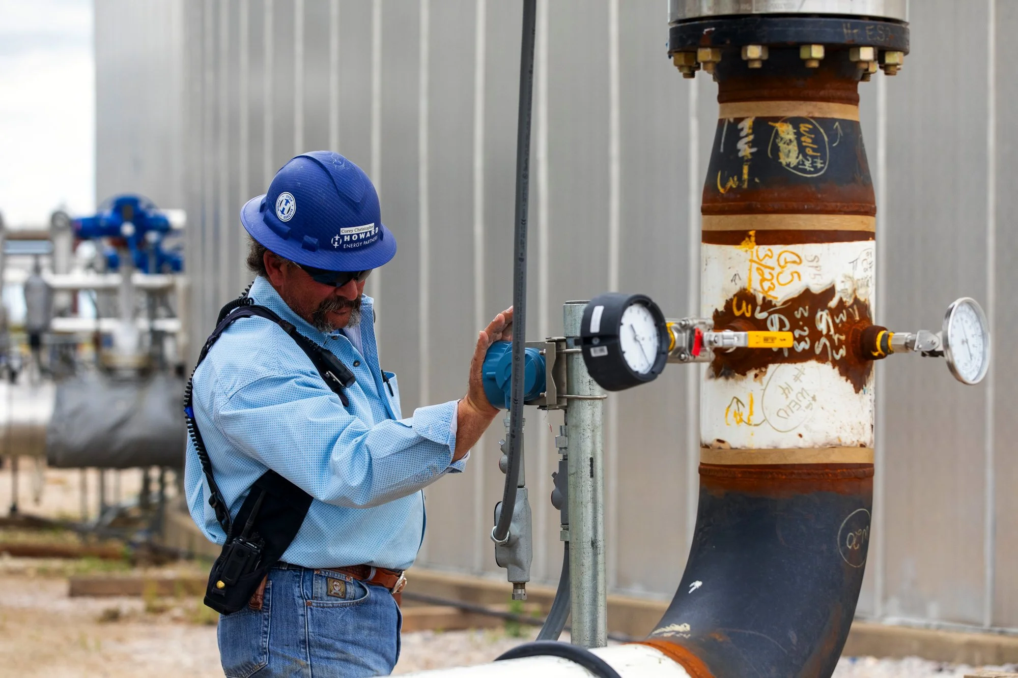 A worker in a blue hard hat and safety glasses inspects a pipeline with pressure gauges at an industrial site.