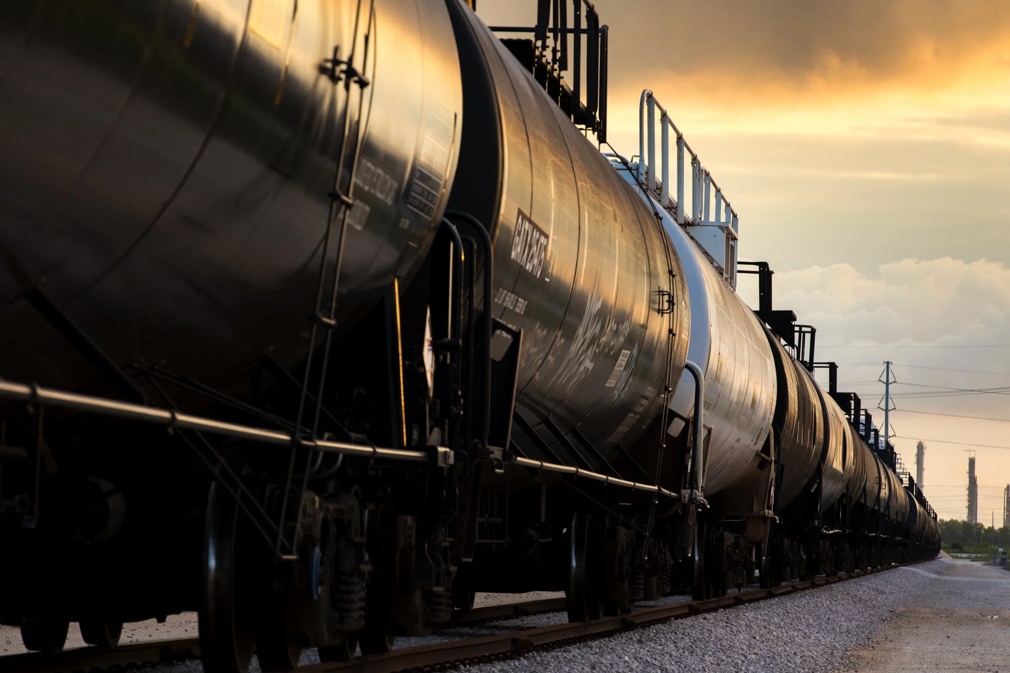 A line of black tanker train cars on railroad tracks at sunset with a sky of orange and yellow clouds in the background.