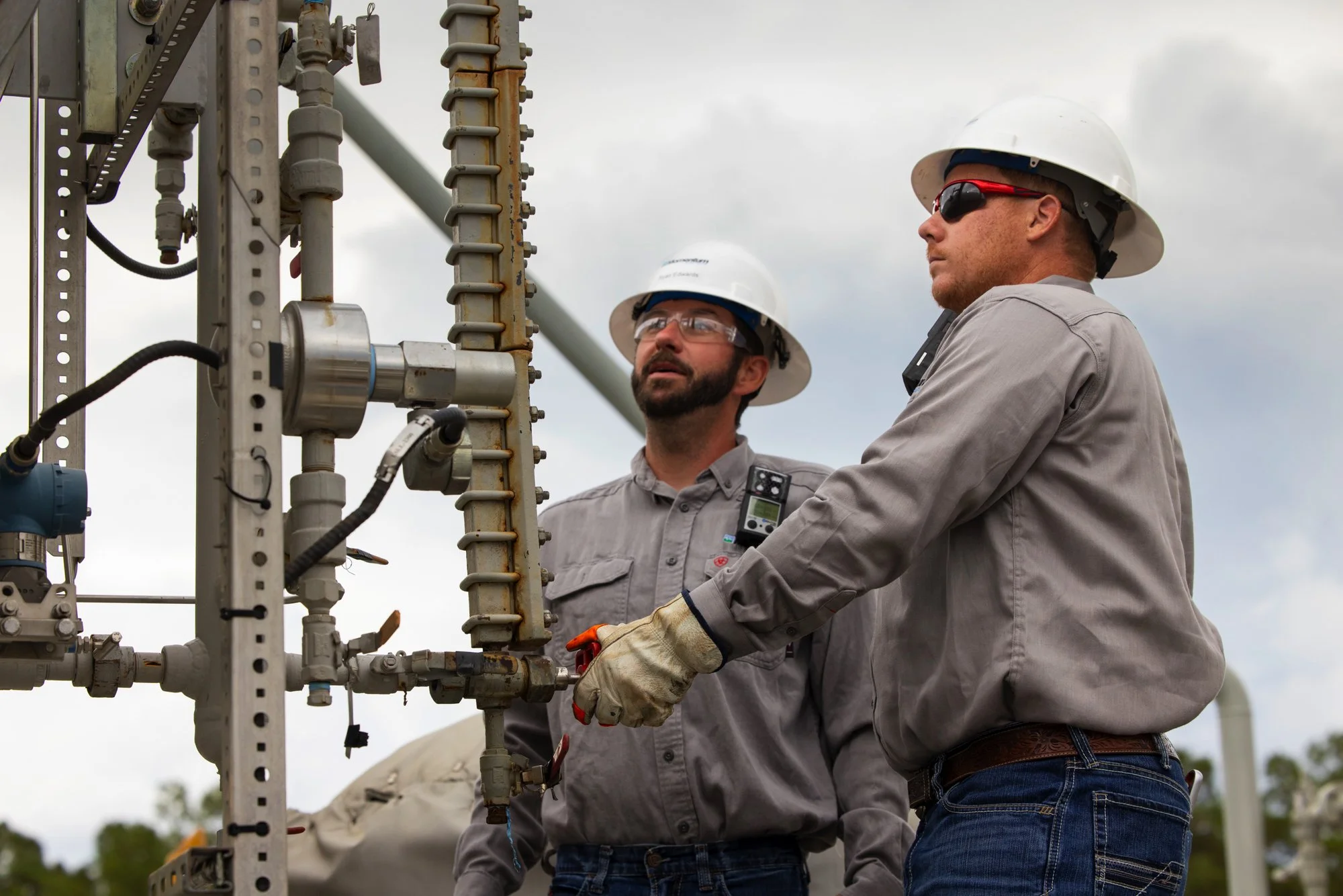 Two male workers wearing hard hats and safety glasses working on industrial piping outdoors.