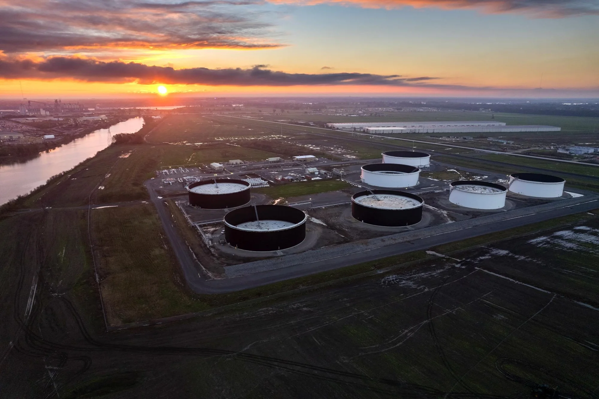 Aerial view of a petroleum storage tank farm at sunset, with multiple black and white cylindrical tanks and a river in the background.
