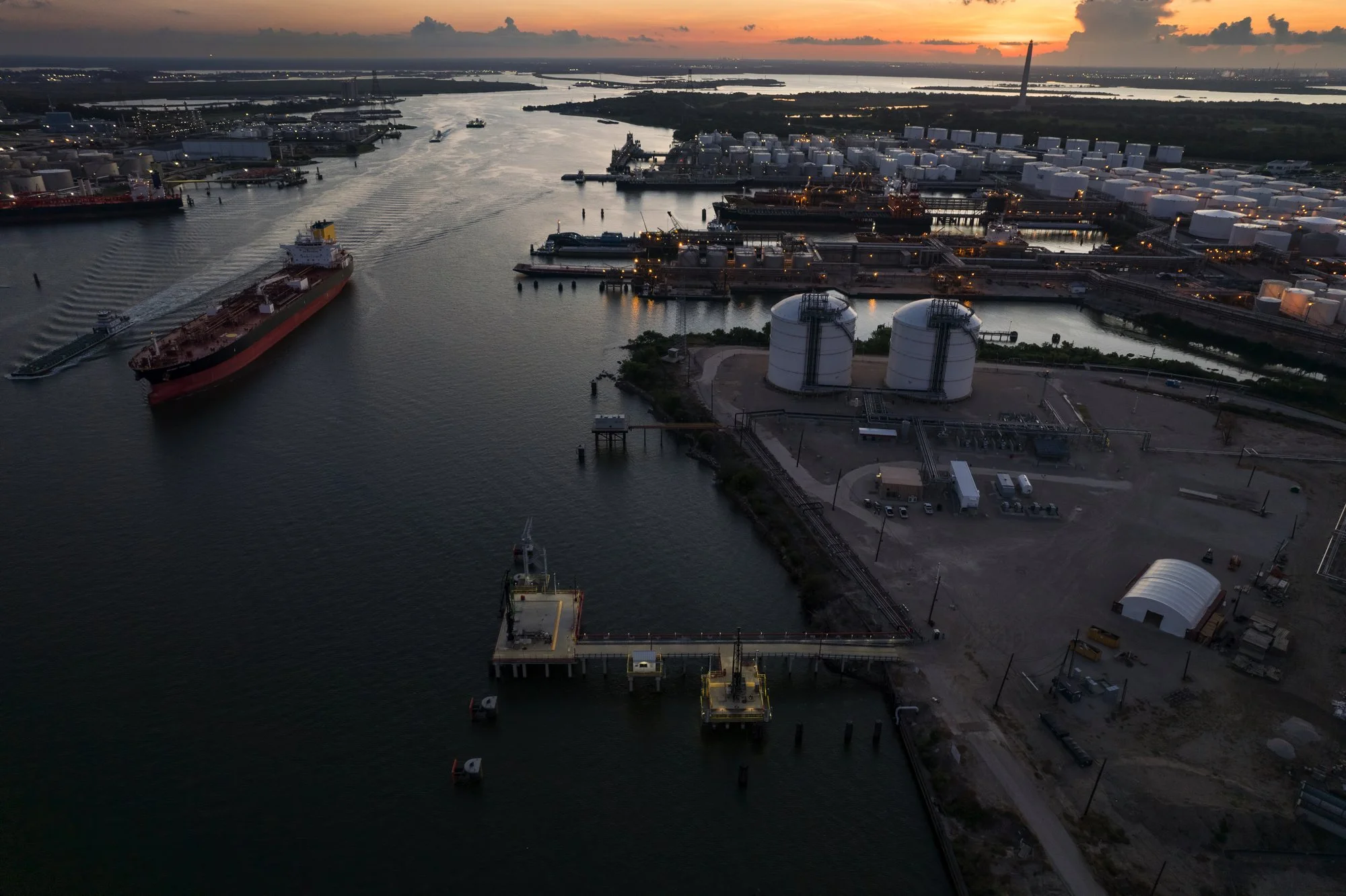 Aerial view of an industrial port at sunset with shipping vessels, storage tanks, warehouses, and refinery facilities along the water.