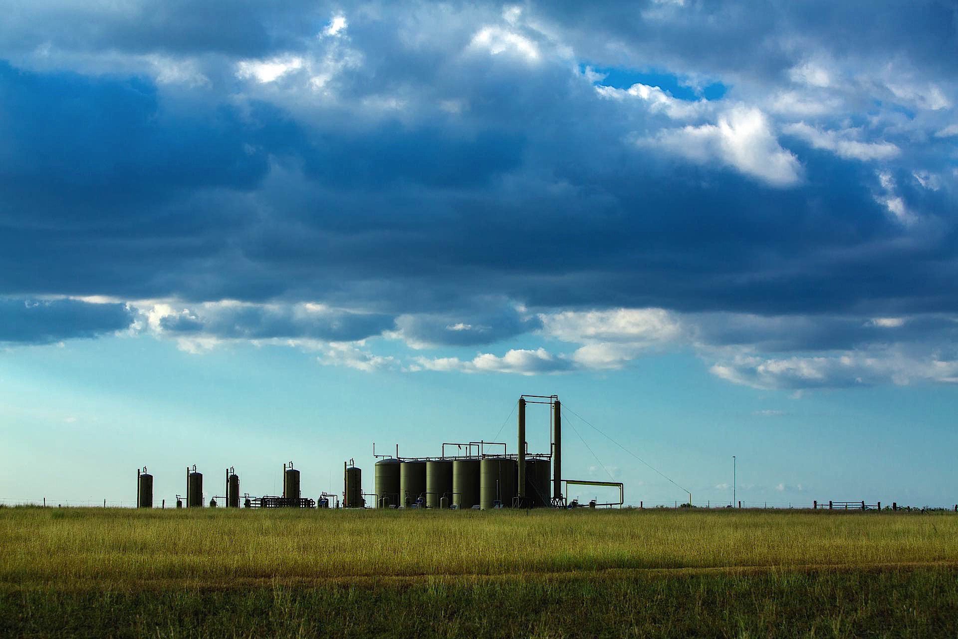 An industrial oil or chemical storage facility with multiple tanks on a grassy plain under a cloudy sky.