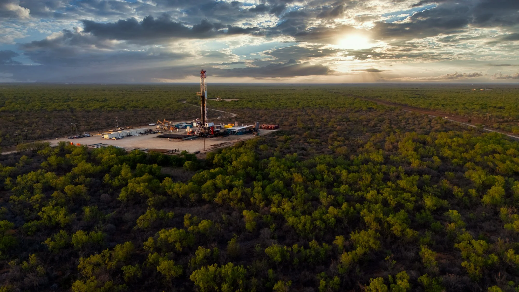 An oil drilling rig surrounded by sparse green shrubbery in a vast, flat landscape during sunset.