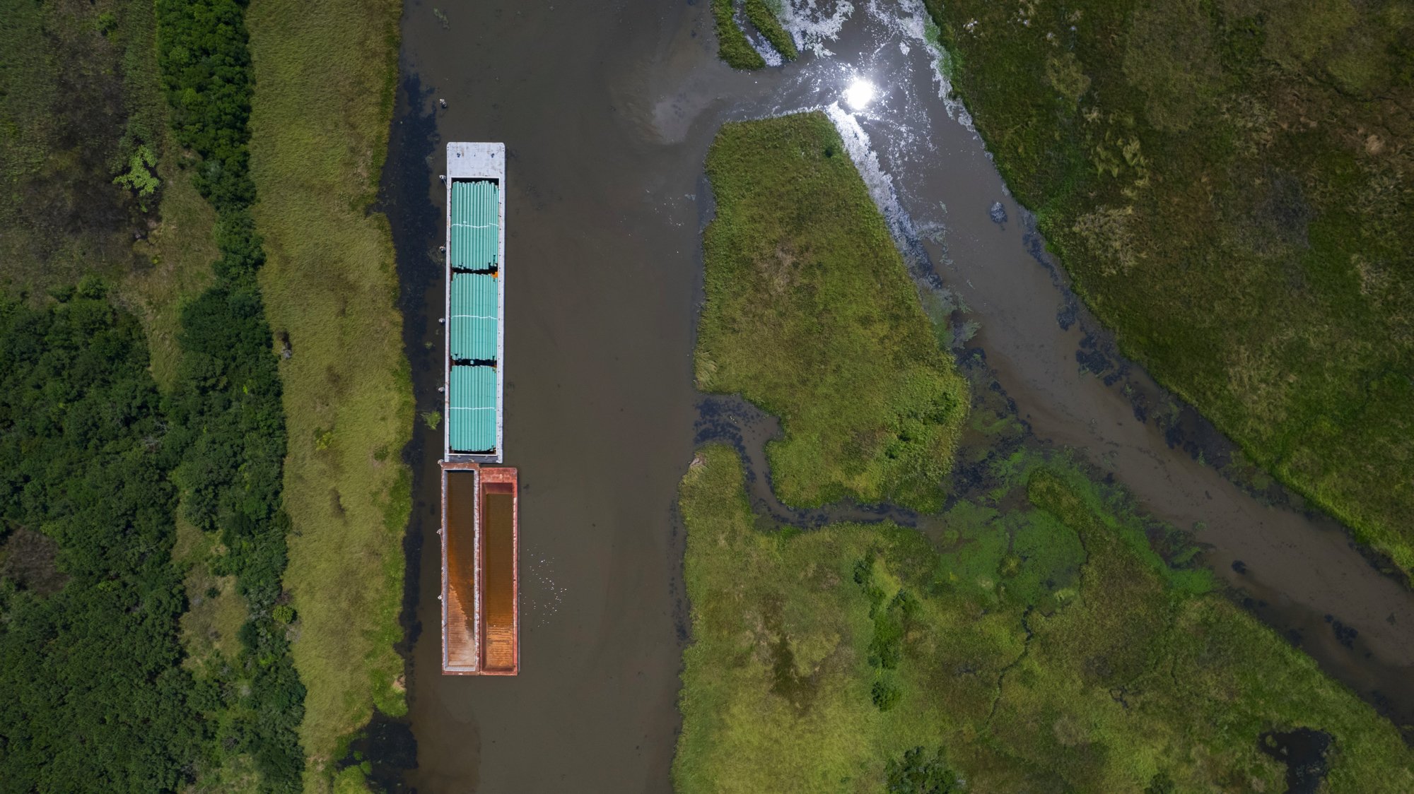 An aerial view of a river with a barge and a tugboat navigating through green marshland.