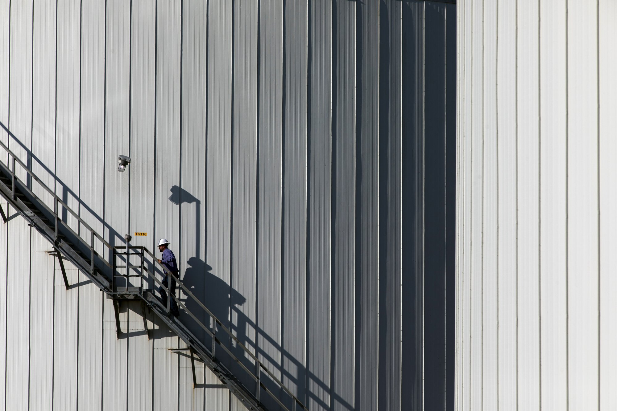 A worker wearing a white safety helmet and blue uniform climbing an external metal staircase attached to a large industrial building with vertical metal siding. The building's wall is gray with vertical panels, and the worker's shadow is cast on the 
