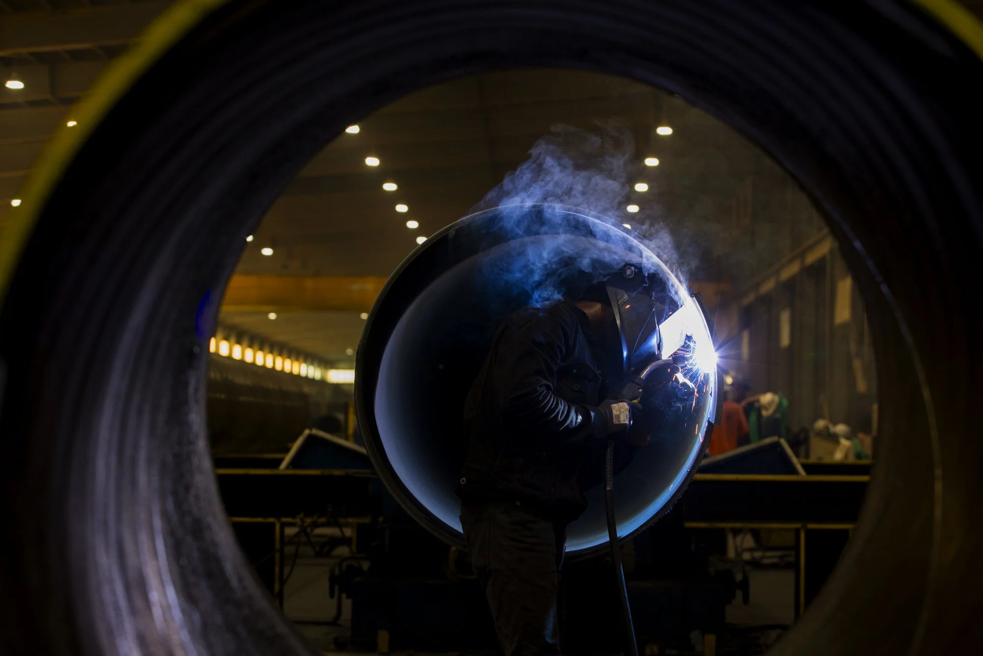 Welder in a dark industrial workshop welding inside a large metal pipe, with visible sparks and smoke.