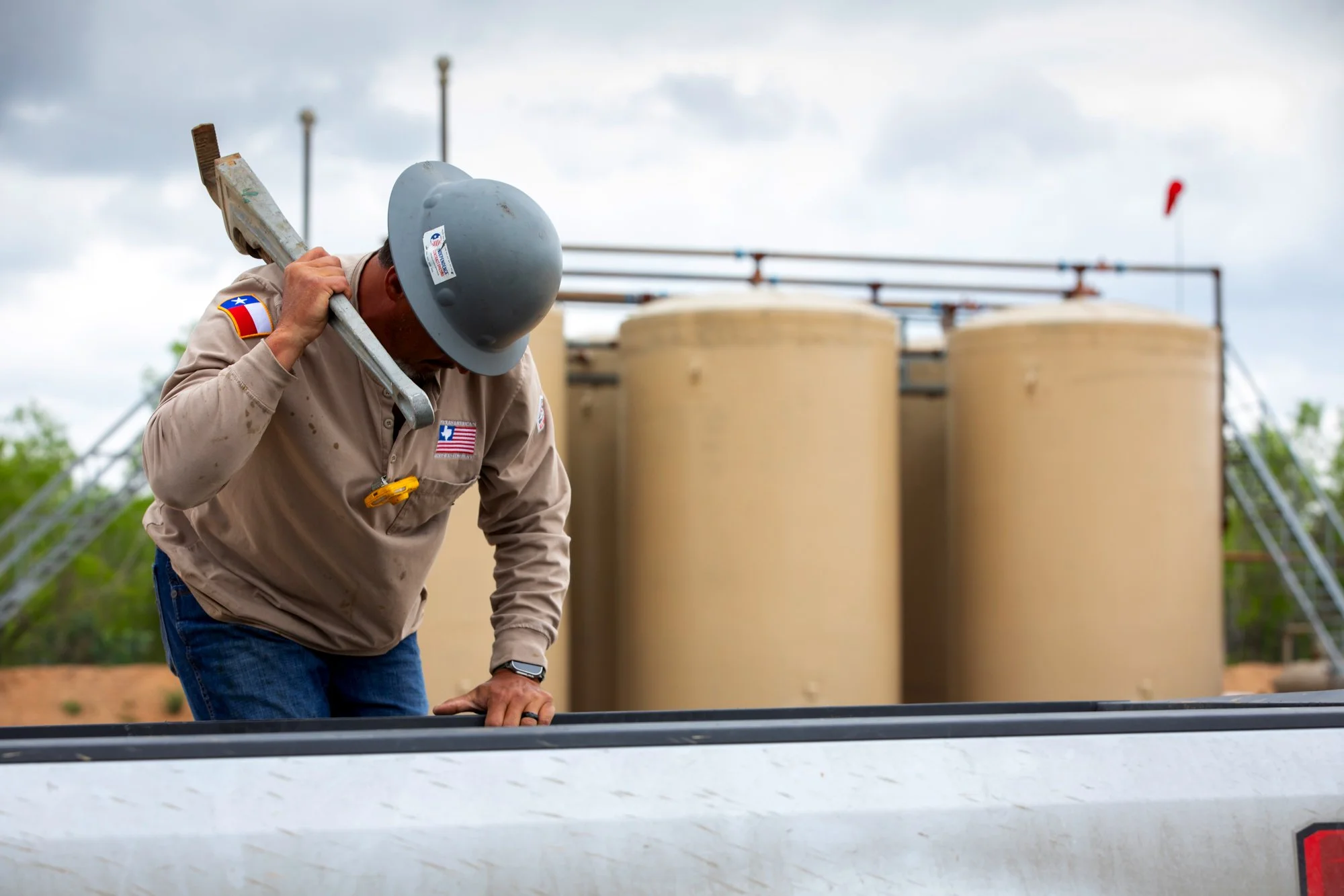 A worker wearing a gray helmet and beige uniform with an American flag patch looks down while holding a tool over a large metal tank or container during daytime.