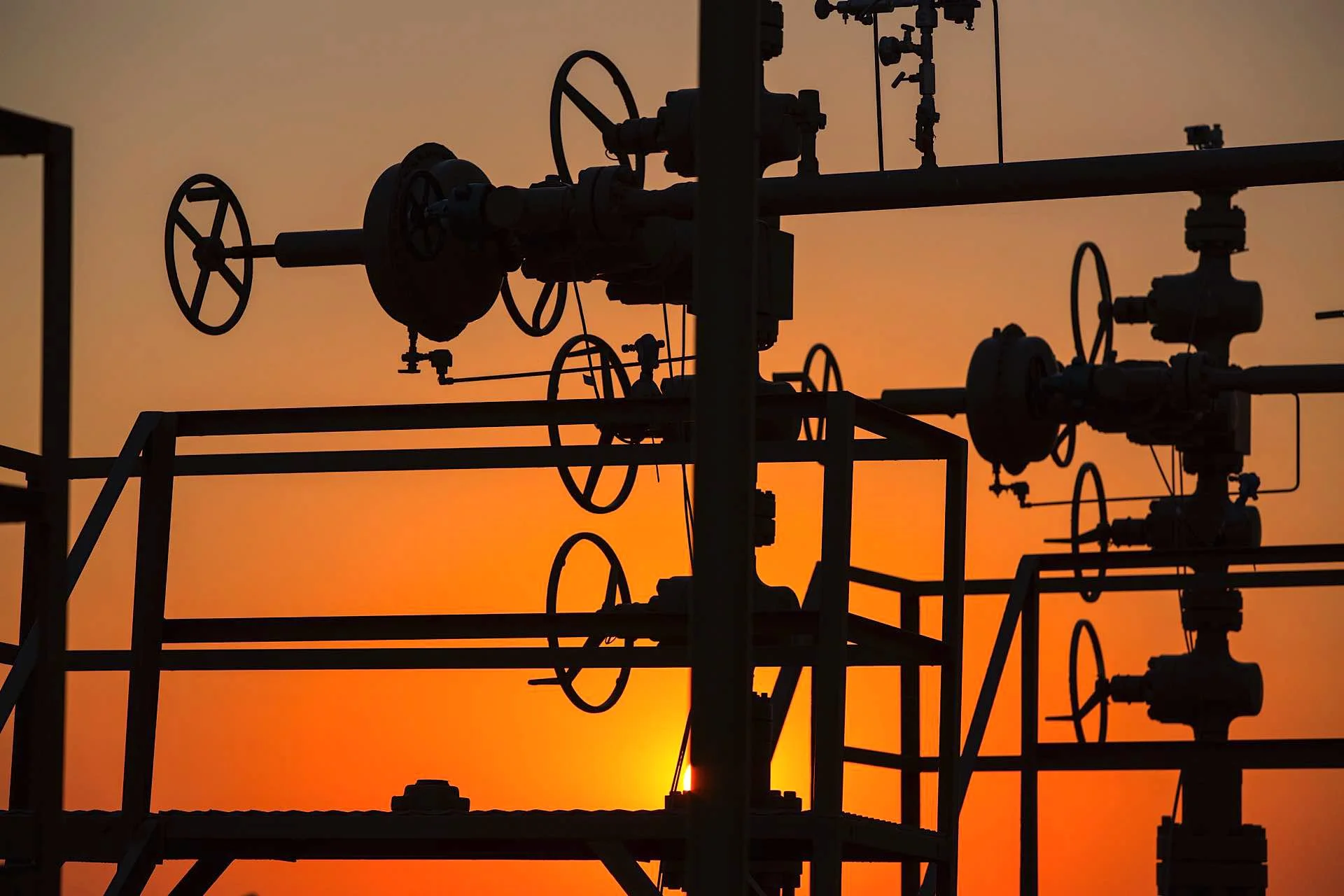 Silhouetted industrial pipelines and valves against a sunset sky.