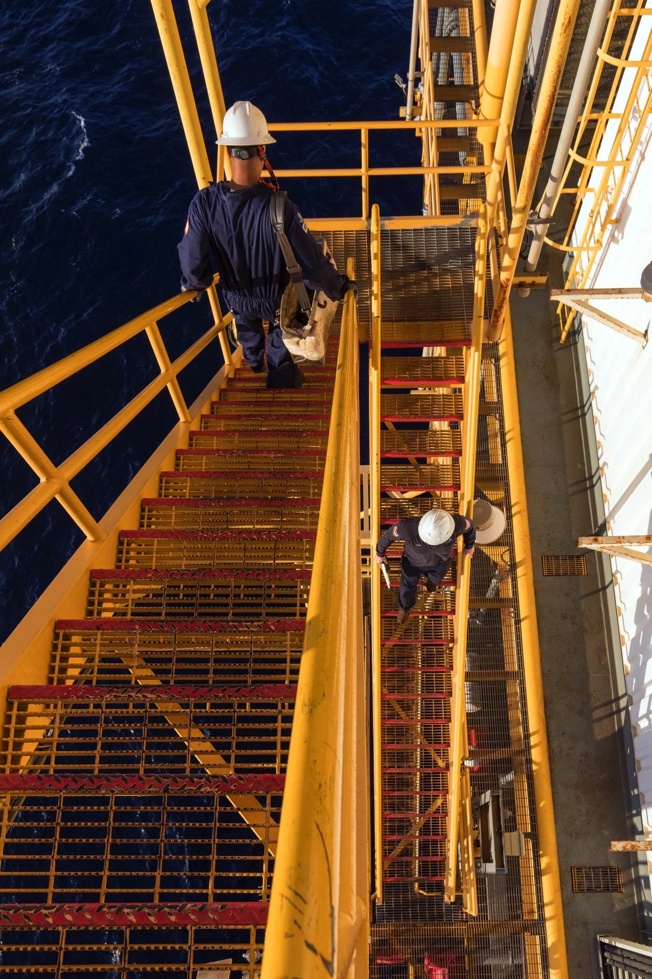 Two workers on a yellow metal staircase on an offshore oil platform, wearing safety helmets and work attire, with ocean water visible below.