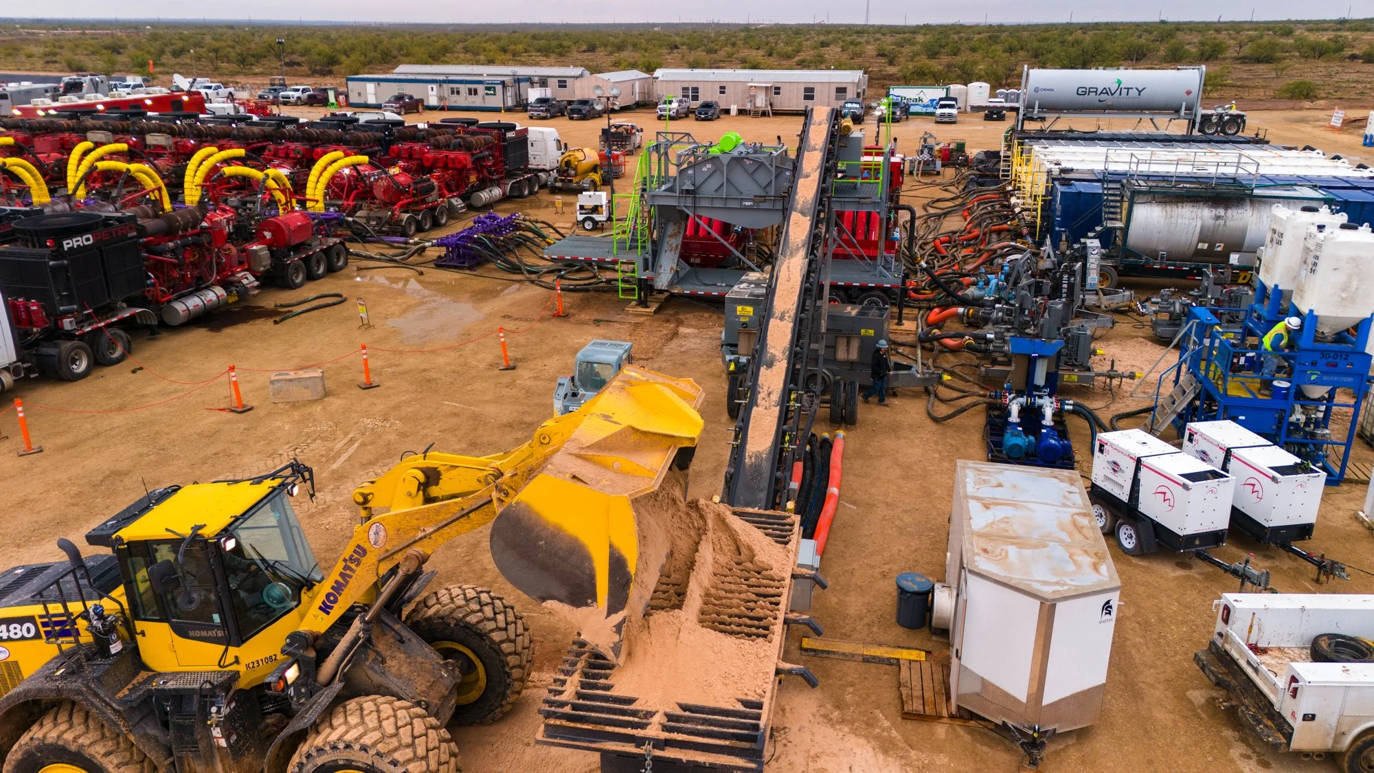 Construction site with a yellow Komatsu front loader excavating sand, surrounded by various industrial equipment, trucks, and machinery in a desert environment.