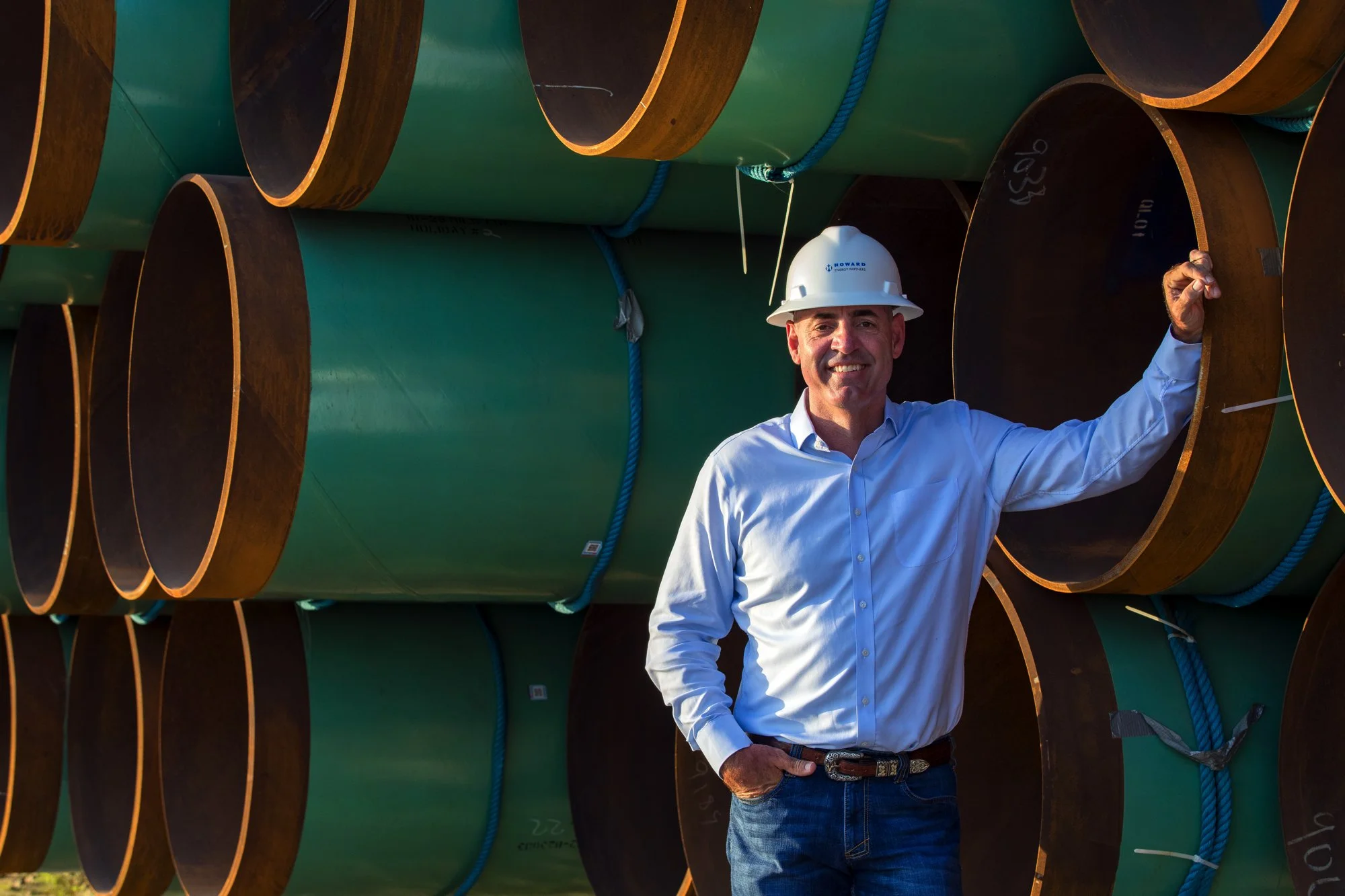 A man in a white hard hat and light blue shirt stands in front of large stacked green and brown pipes, smiling at the camera.