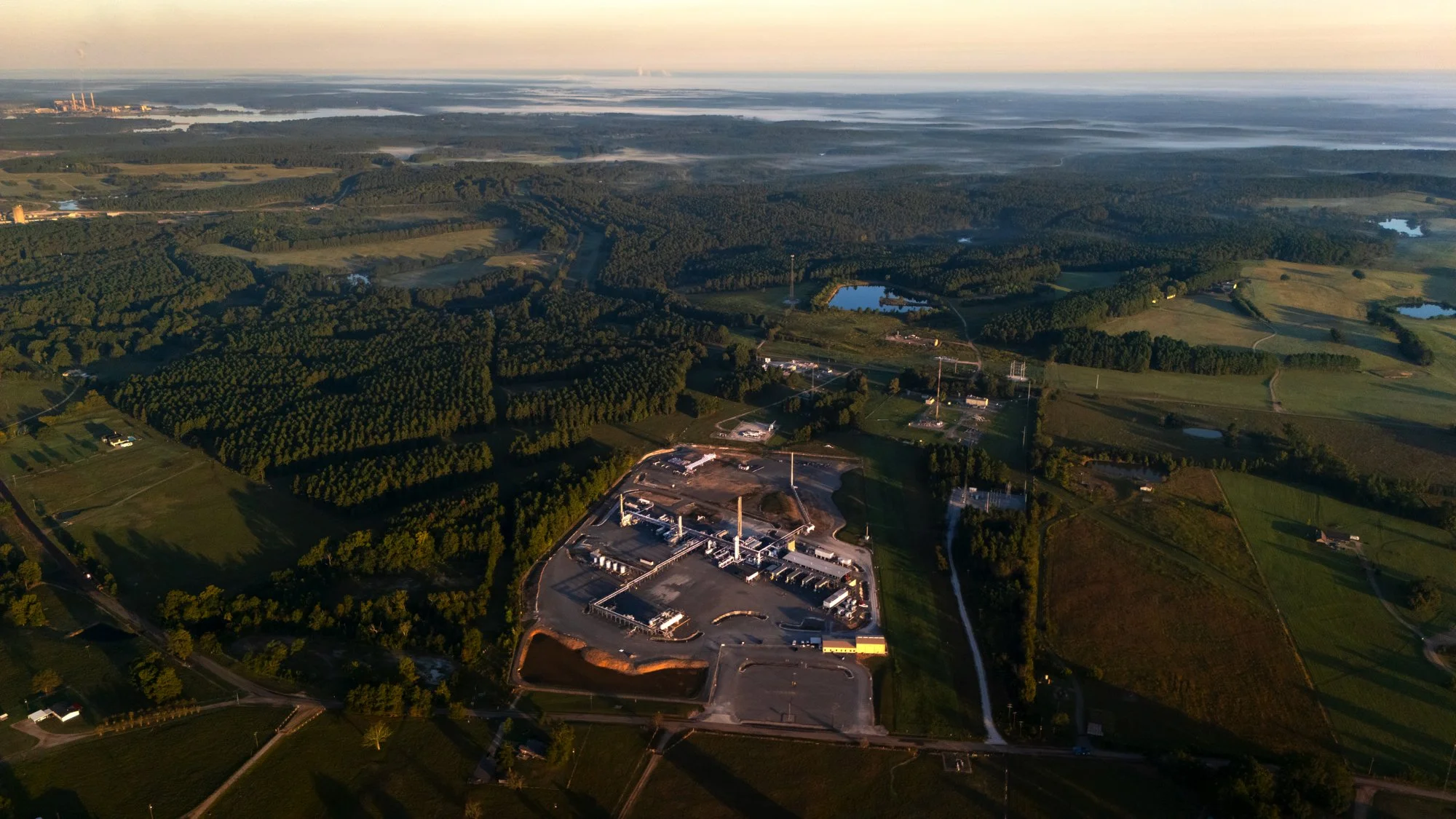 Aerial view of a rural landscape with fields, trees, lakes, and a power plant in the distance. The foreground shows a construction site or industrial facility.
