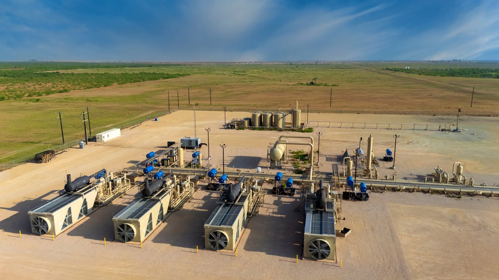 Aerial view of an industrial facility with large machinery, pipes, and storage tanks in a desert area, with open fields and clear blue sky in the background.