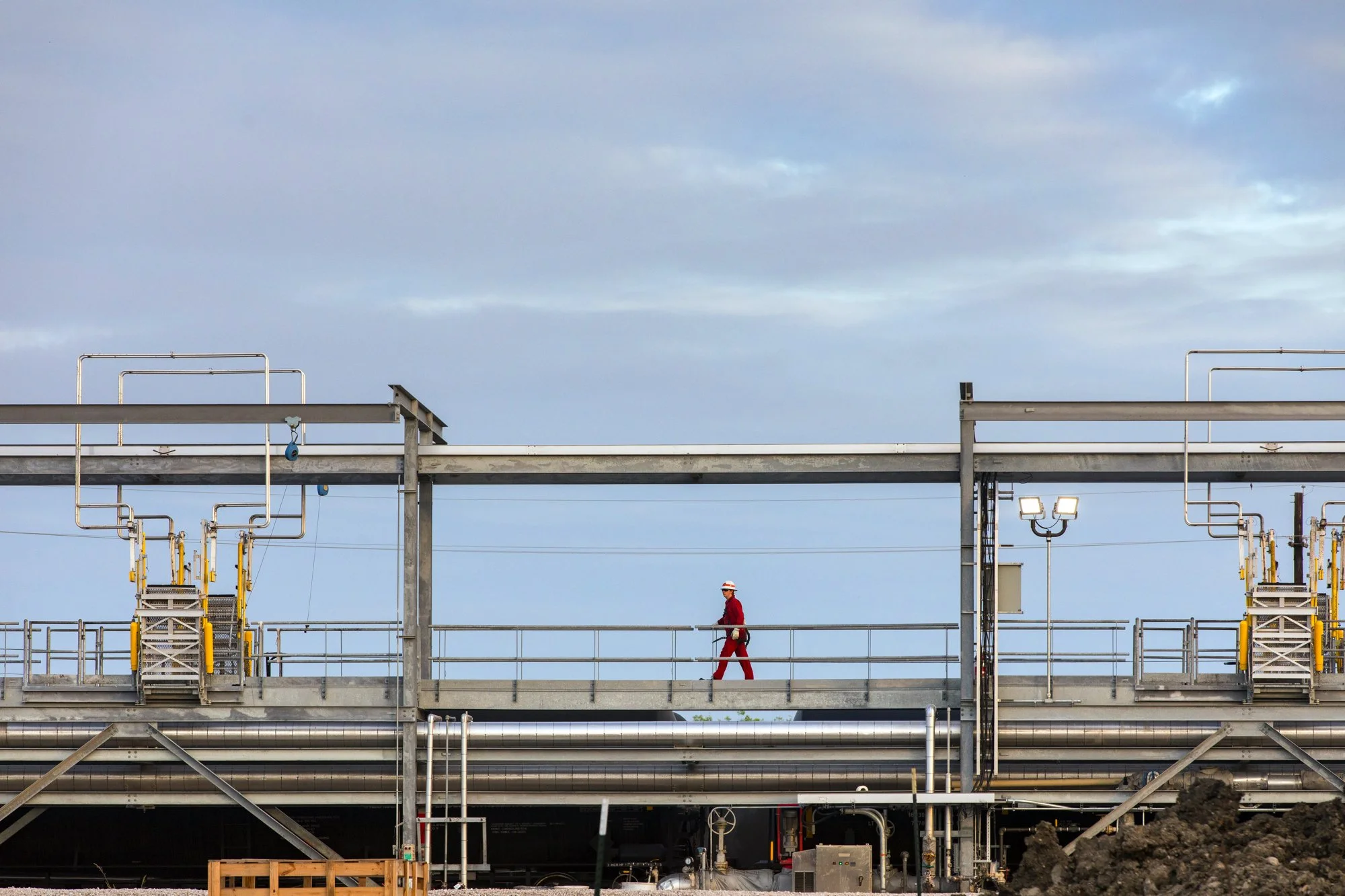 A worker in red overalls and a white hard hat walking on an industrial platform with pipes and equipment under a cloudy sky.