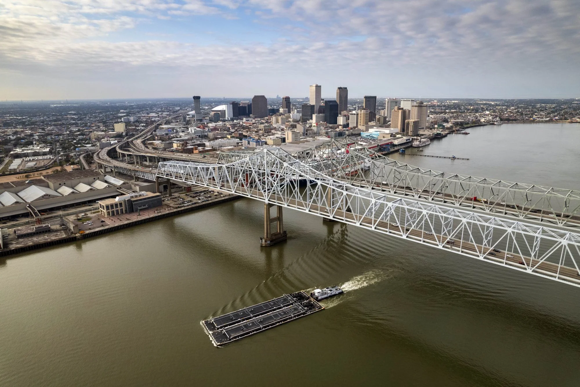 Aerial view of a city skyline with a large bridge over a river, a boat on the water, and various buildings and highways, during daytime with cloudy sky.
