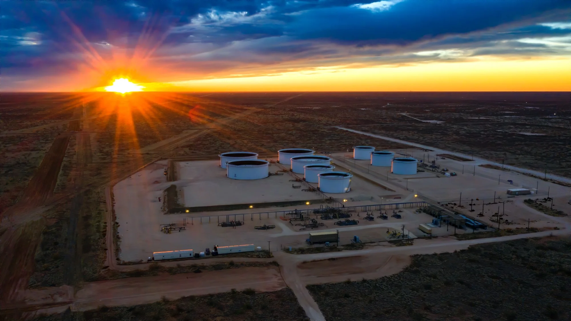 Aerial view of a large fuel storage facility with multiple white tanks during sunset in a desert landscape.