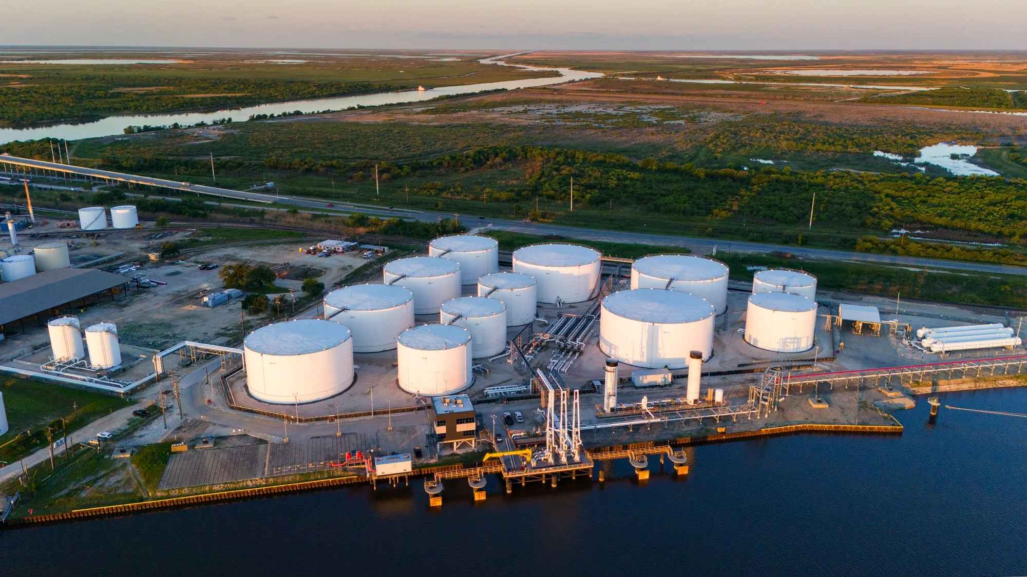 Aerial view of a large industrial storage tank facility next to a body of water, with pipelines and vehicles, surrounded by green fields and wetlands.