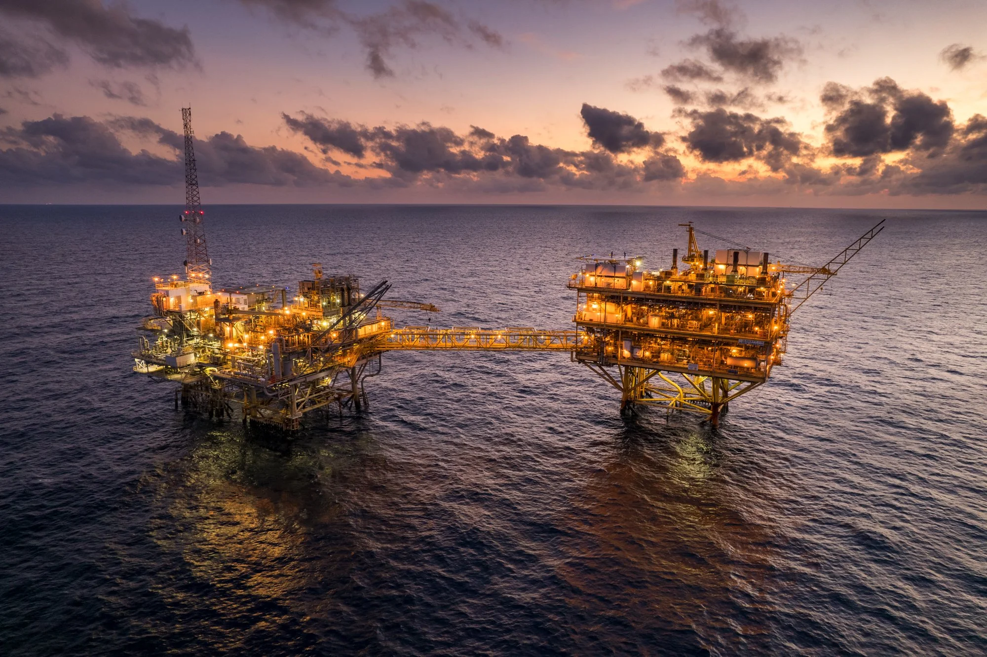 An offshore oil rig with illuminated structures in the ocean at sunset, with a cloudy sky in the background.