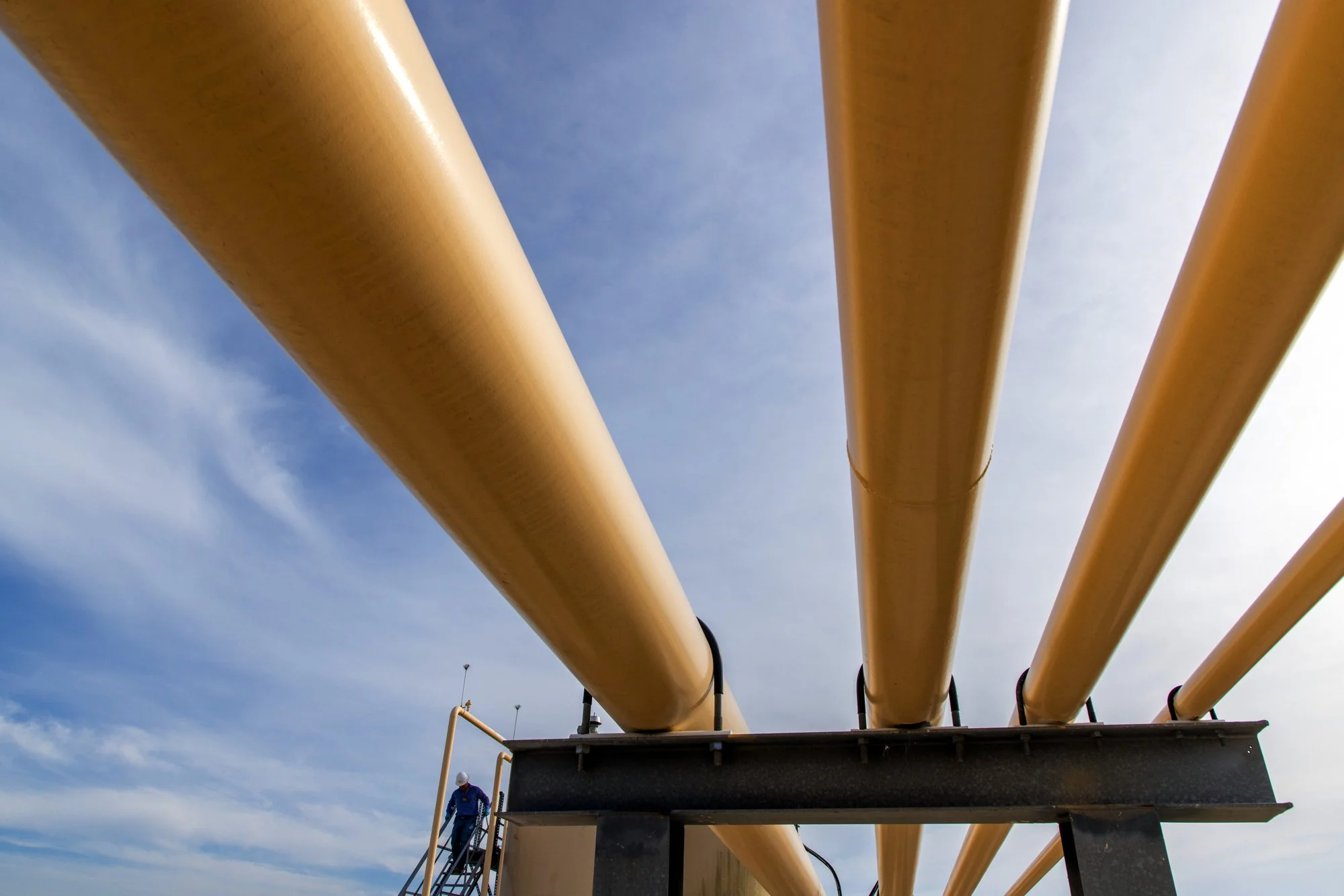 A worker wearing a blue jumpsuit and a helmet walking on a metal platform beneath three large yellow pipelines against a partly cloudy sky.