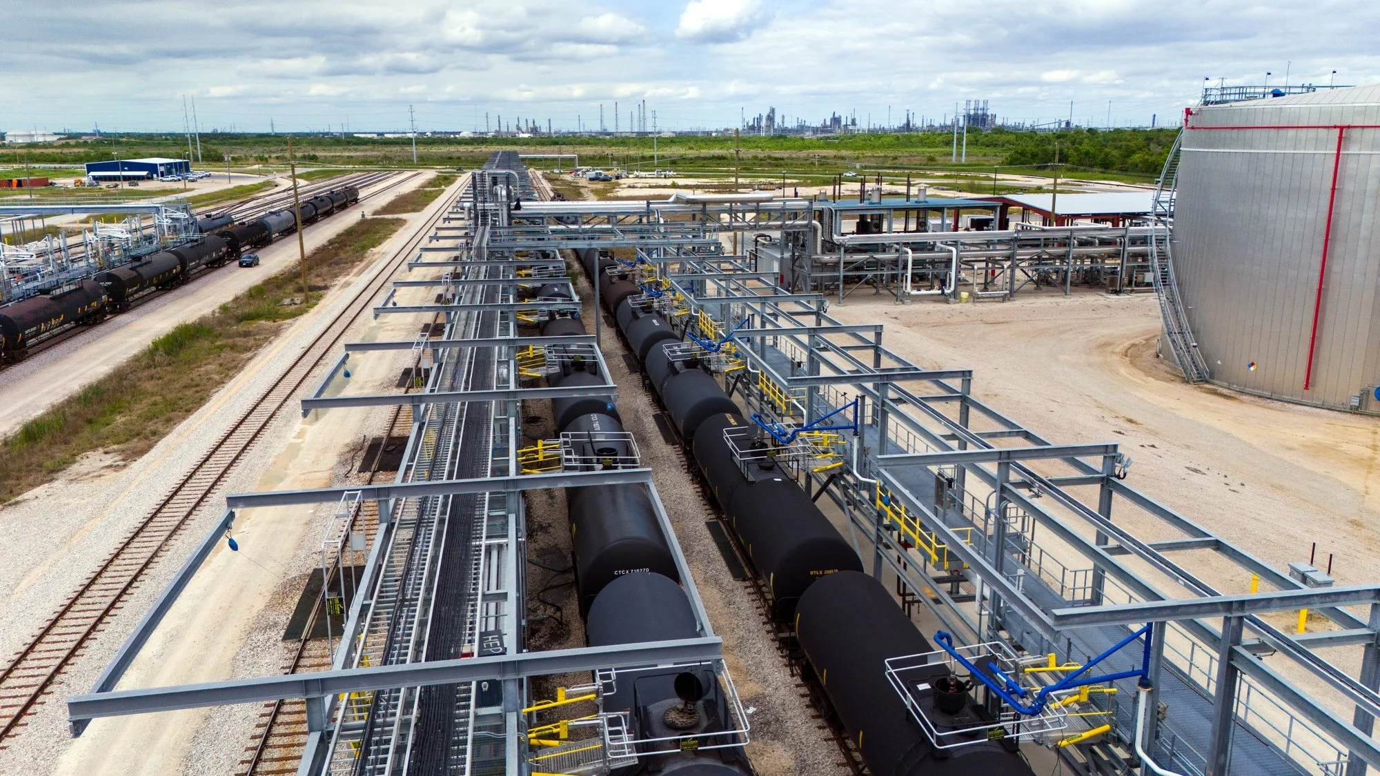 View of an industrial facility with train tracks, black tanker railcars, and extensive piping and metal structures, with a city skyline in the distance under a cloudy sky.