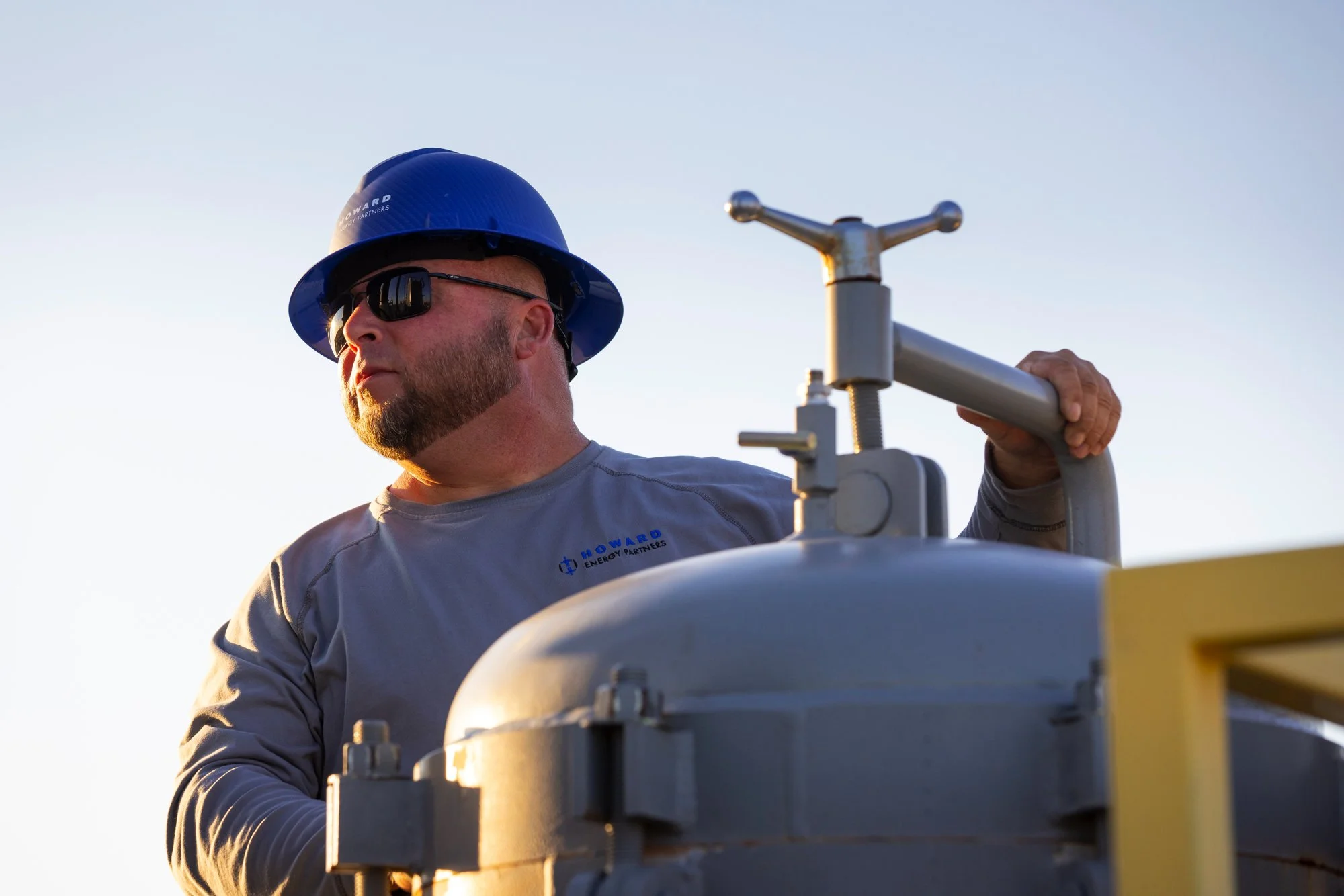 A man wearing a blue hard hat and sunglasses operates outdoor industrial equipment against a clear sky.