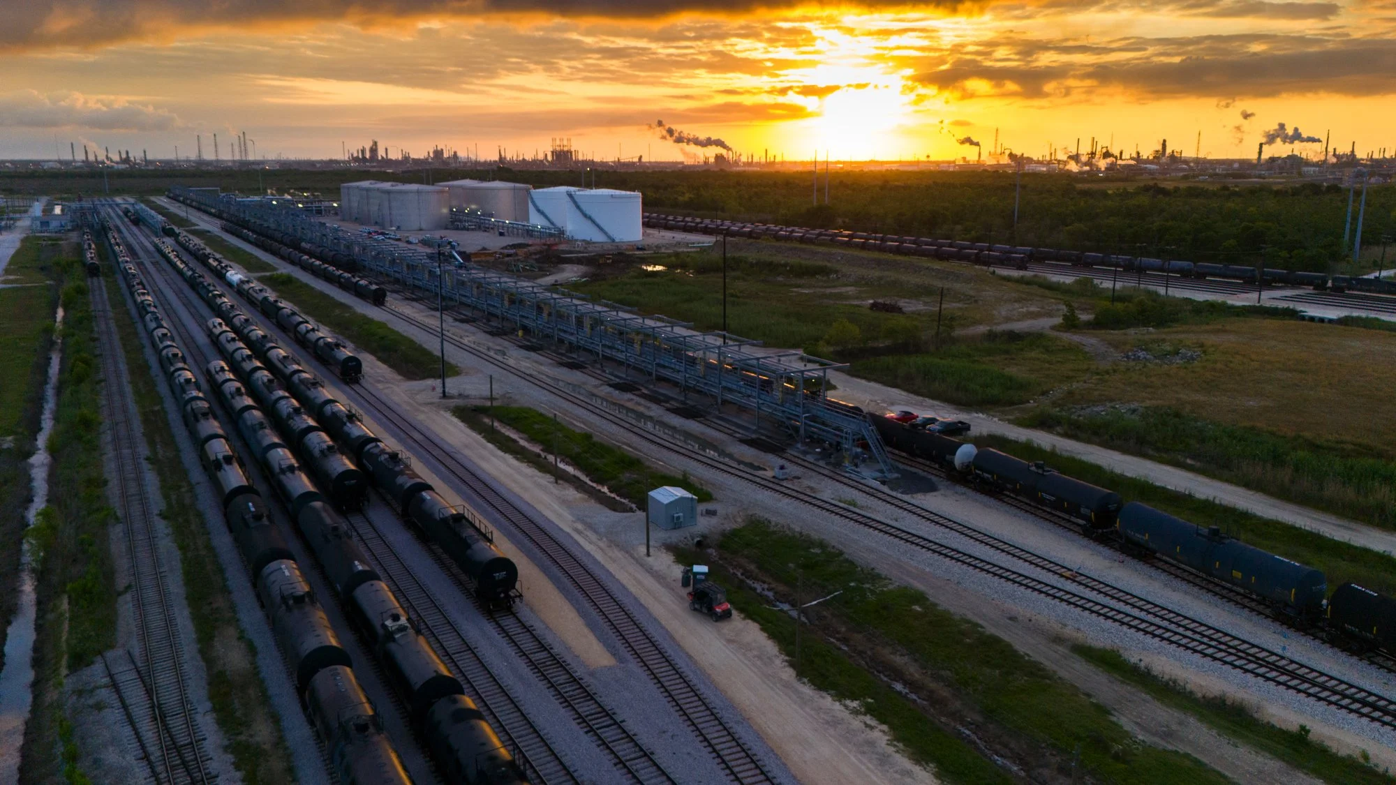 An industrial rail yard at sunset with multiple train tracks, black tanker cars, and large white storage tanks in the background, with smoke and factory emissions visible in the distance.