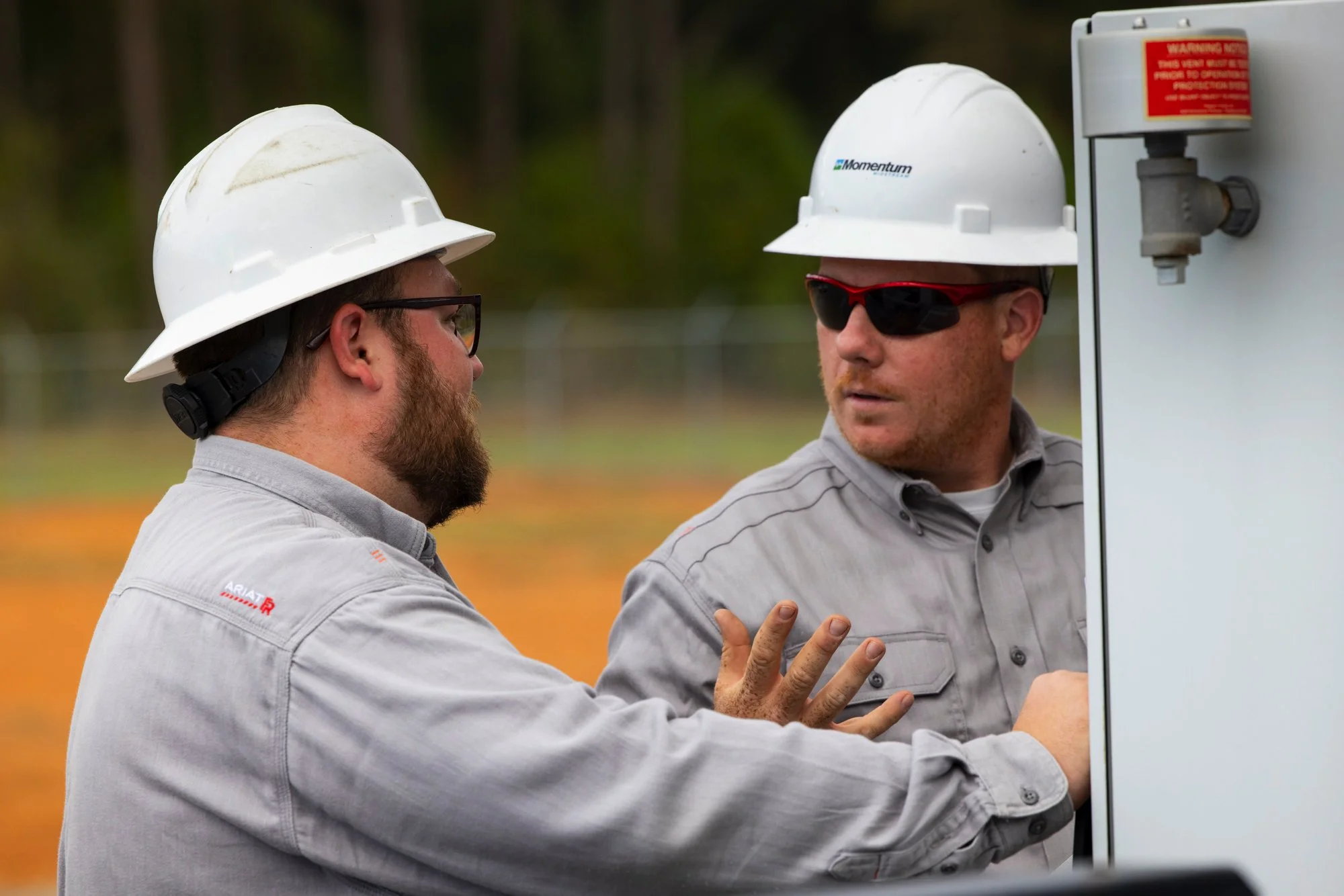 Two male utility workers wearing white safety helmets, gray work shirts, and sunglasses are having a discussion outdoors. One is gesturing towards a piece of equipment on a utility box.