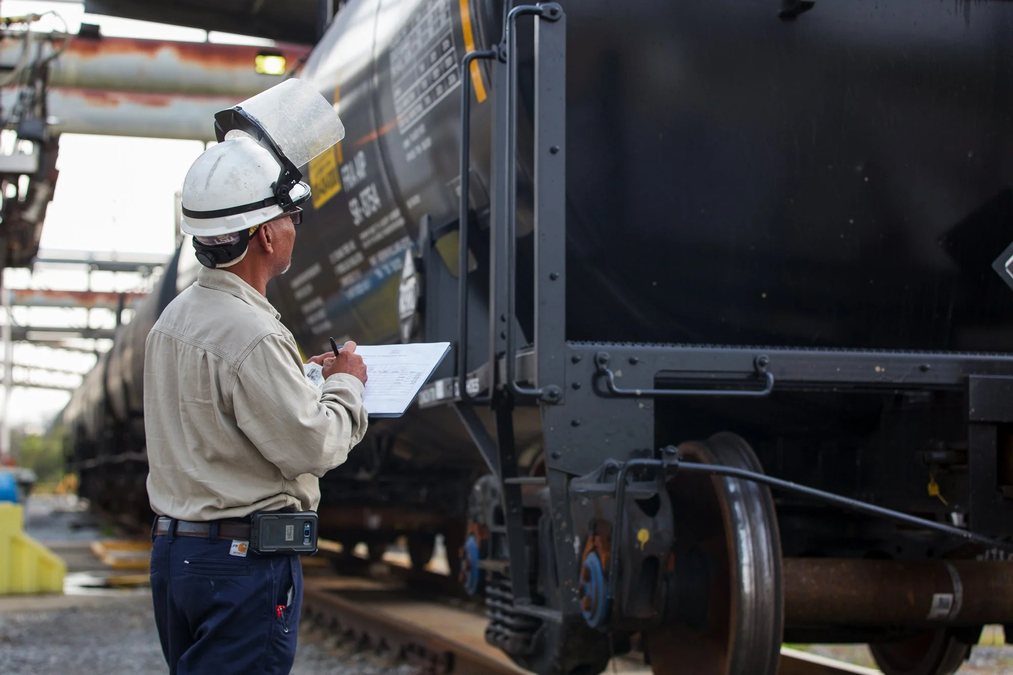 A worker in a hard hat and safety gear inspecting a train car at a rail yard, holding a clipboard and taking notes.
