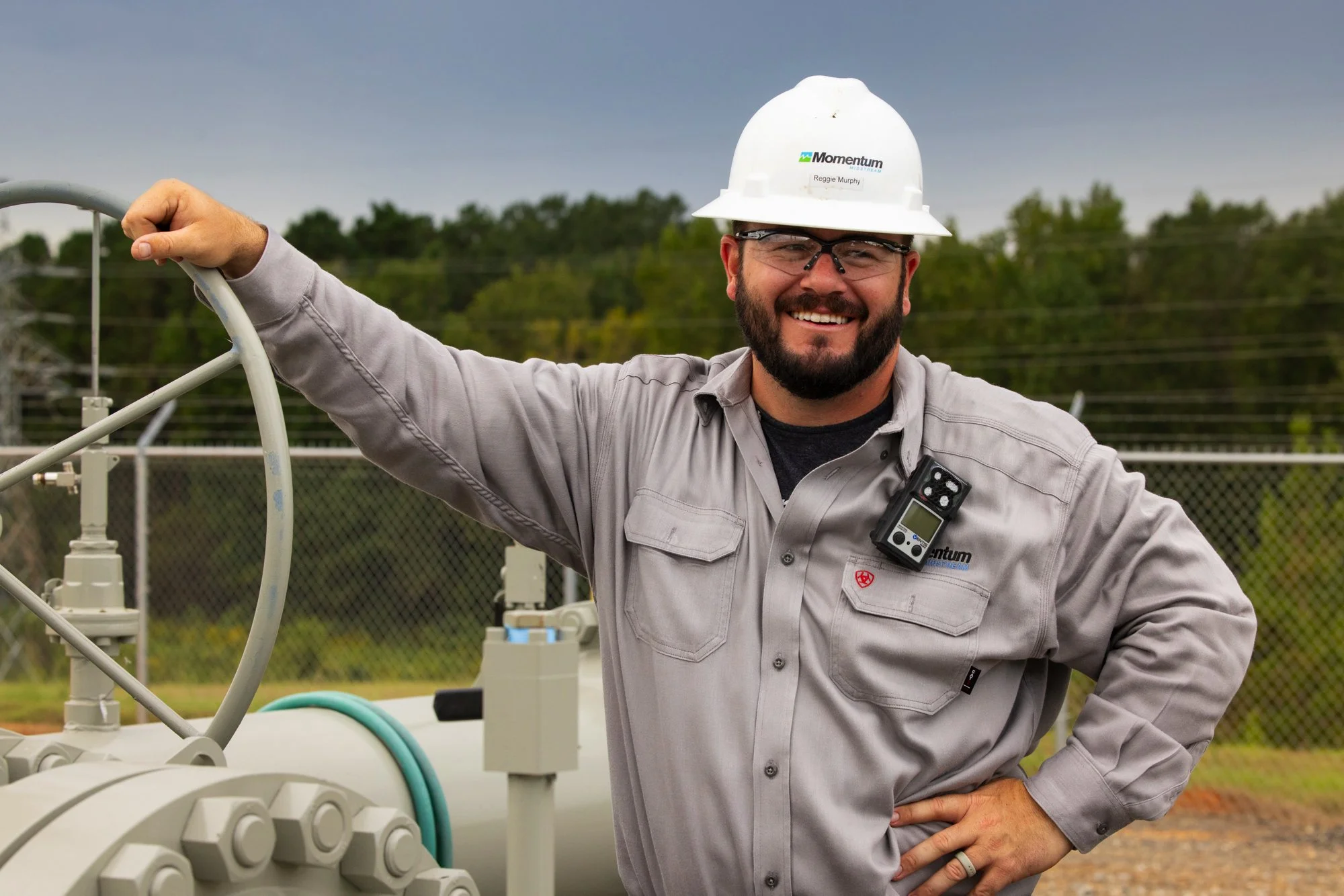 A smiling man wearing a white hard hat, safety glasses, and a gray long-sleeve shirt standing outdoors near industrial equipment and a chain-link fence with trees in the background.
