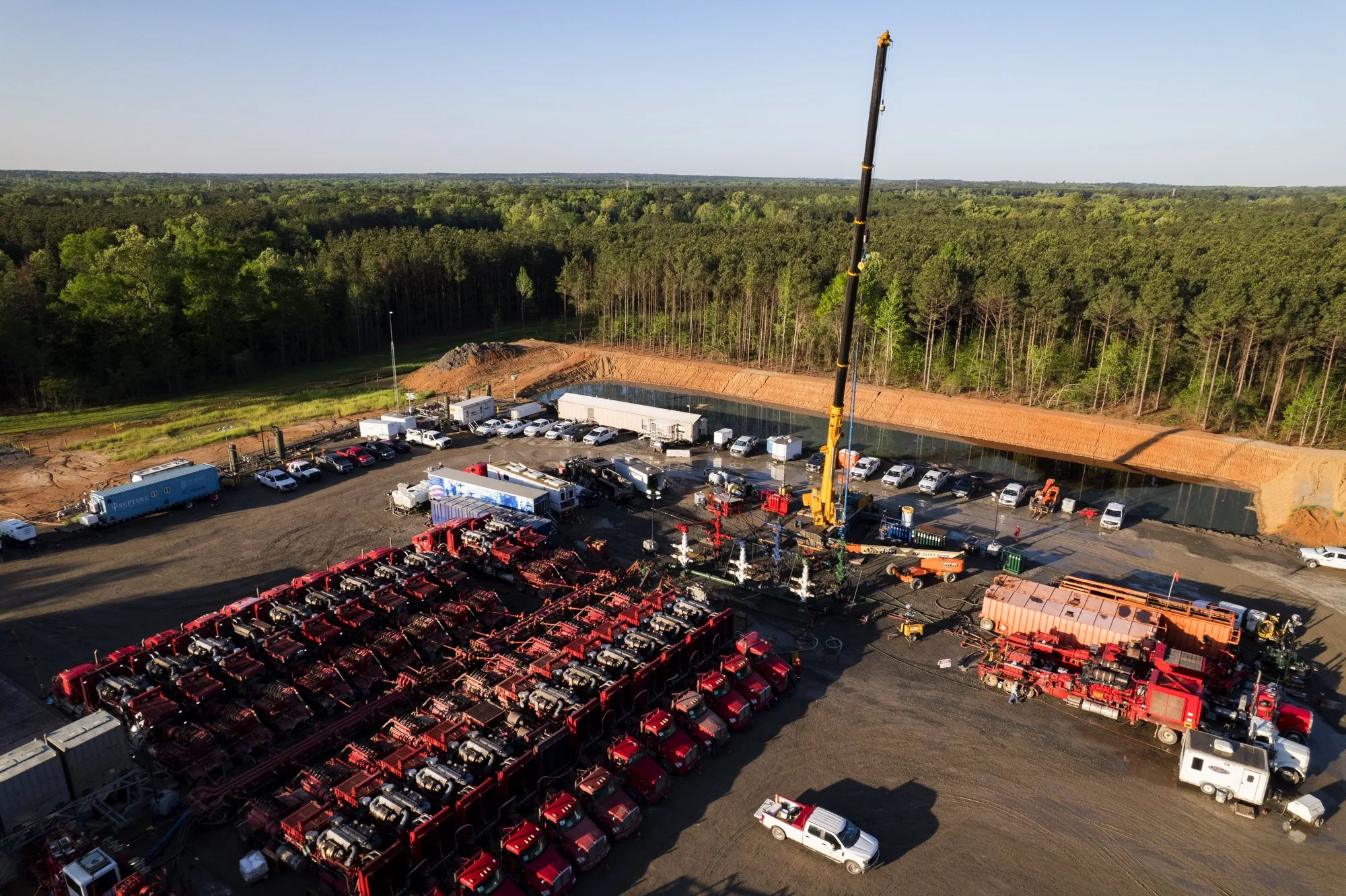 Aerial view of an industrial construction site with red trucks, trucks, construction equipment, and a crane lifting materials, surrounded by forest.