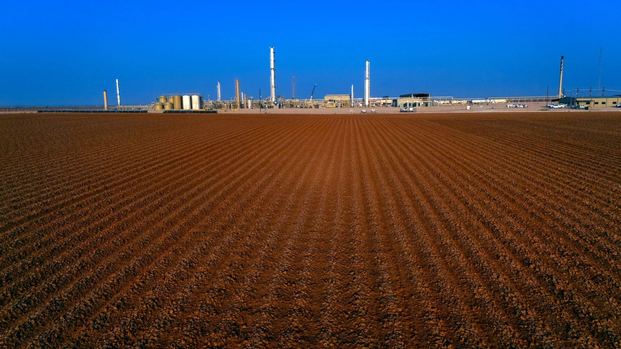 A large, freshly plowed field with evenly spaced rows of soil extends toward an industrial refinery in the distance under a clear blue sky.