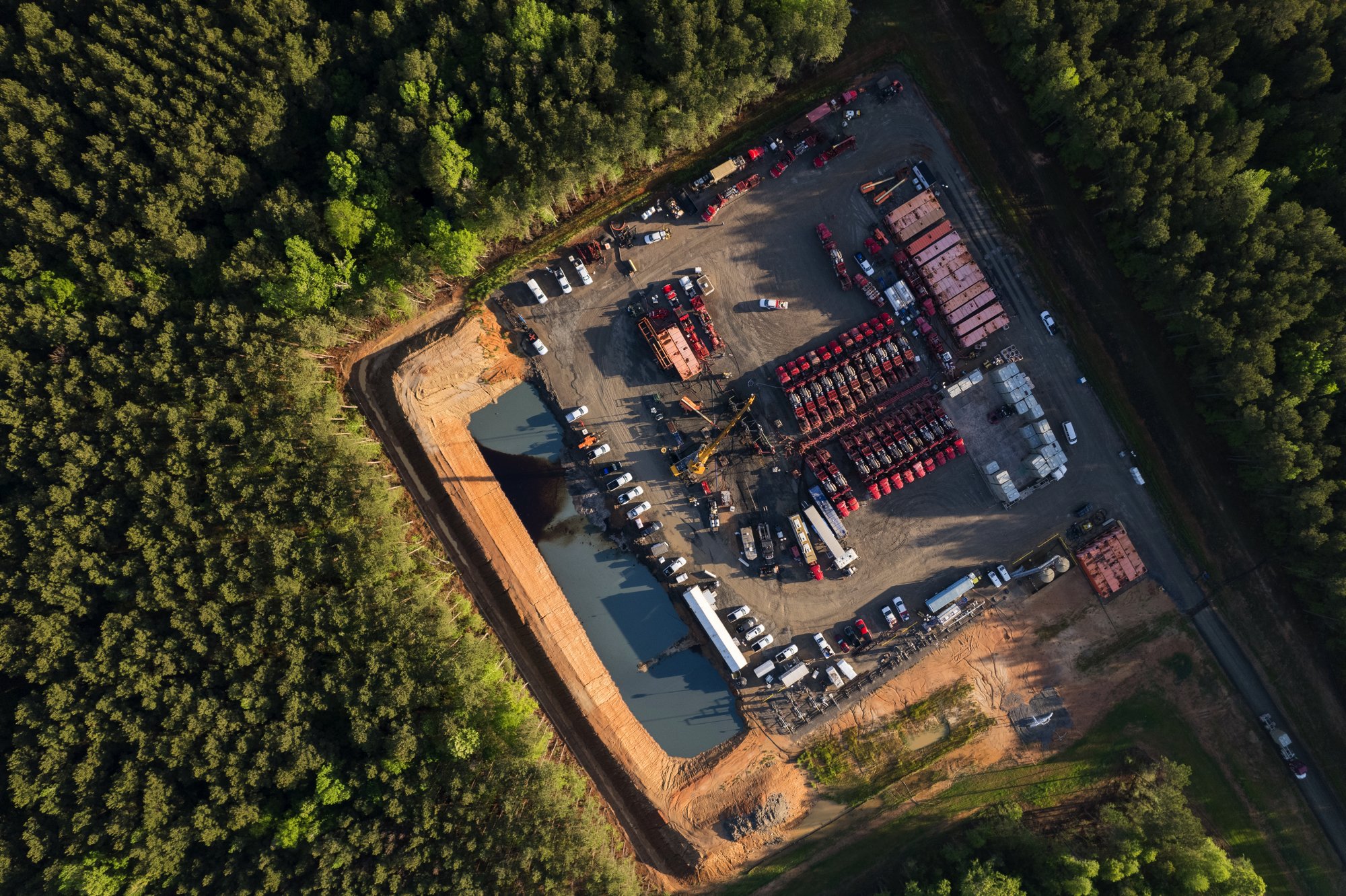 Aerial view of a construction site with a pond, trucks, and construction machinery surrounded by dense trees.