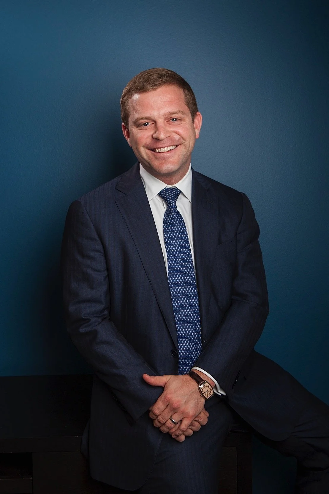A smiling man in a dark gray business suit, white shirt, and blue patterned tie, sitting against a blue wall