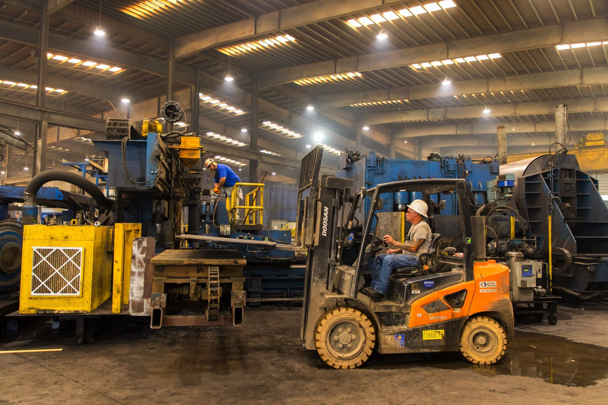 Workers operating heavy machinery in an industrial warehouse setting, with large blue equipment and overhead lighting.