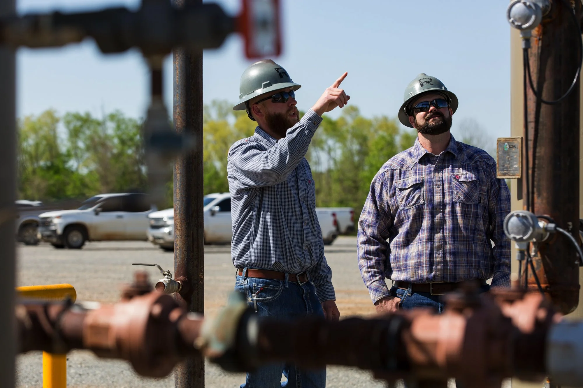 Two men wearing safety helmets and sunglasses inspecting industrial equipment outdoors, with parked cars and trees in the background.
