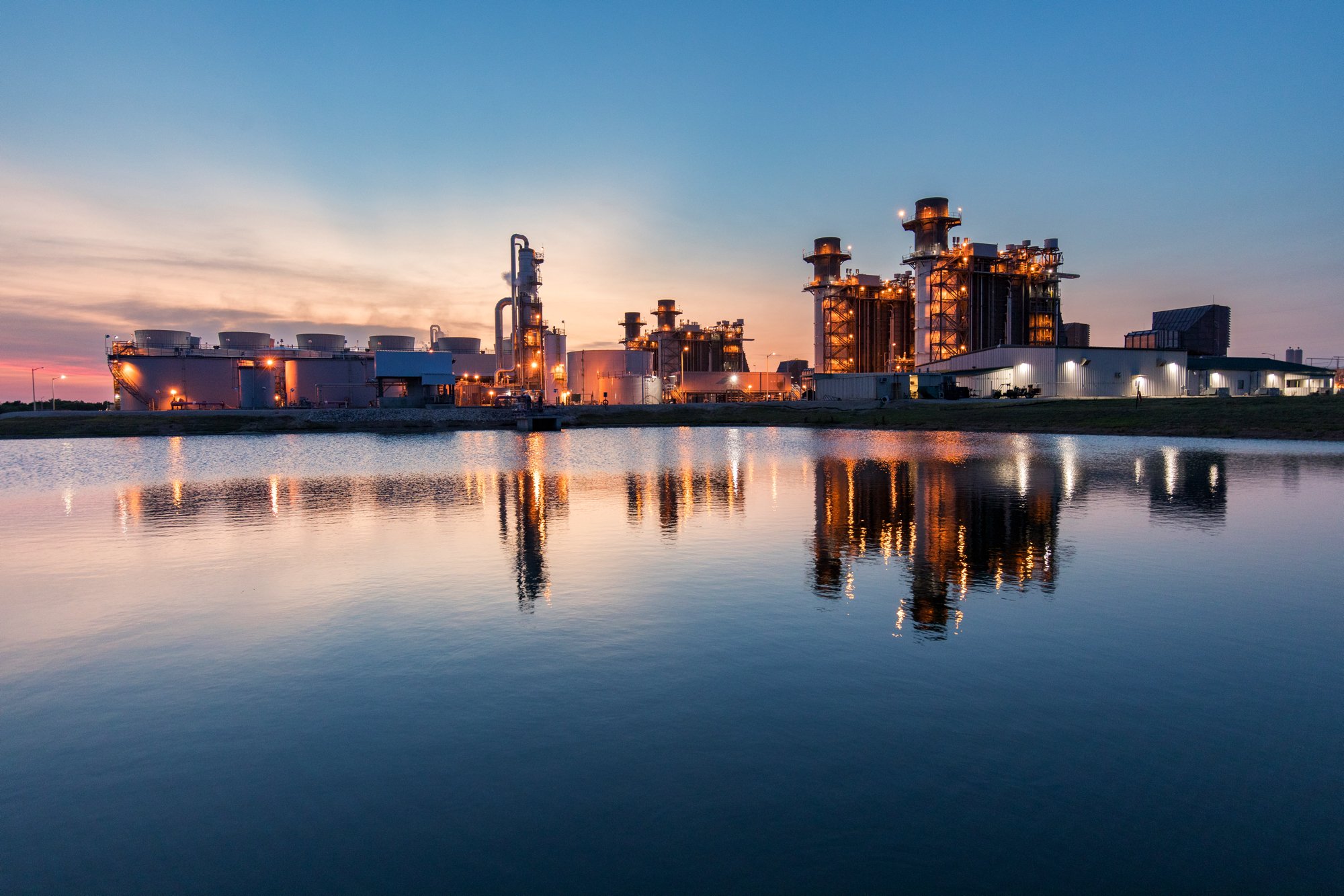 Industrial complex with factories and tall structures reflecting on a body of water at sunset