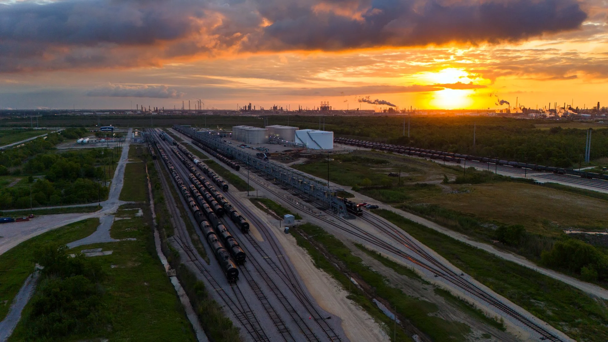 Aerial view of train tracks with freight trains at sunset, industrial factories in the distance, and cloudy sky.