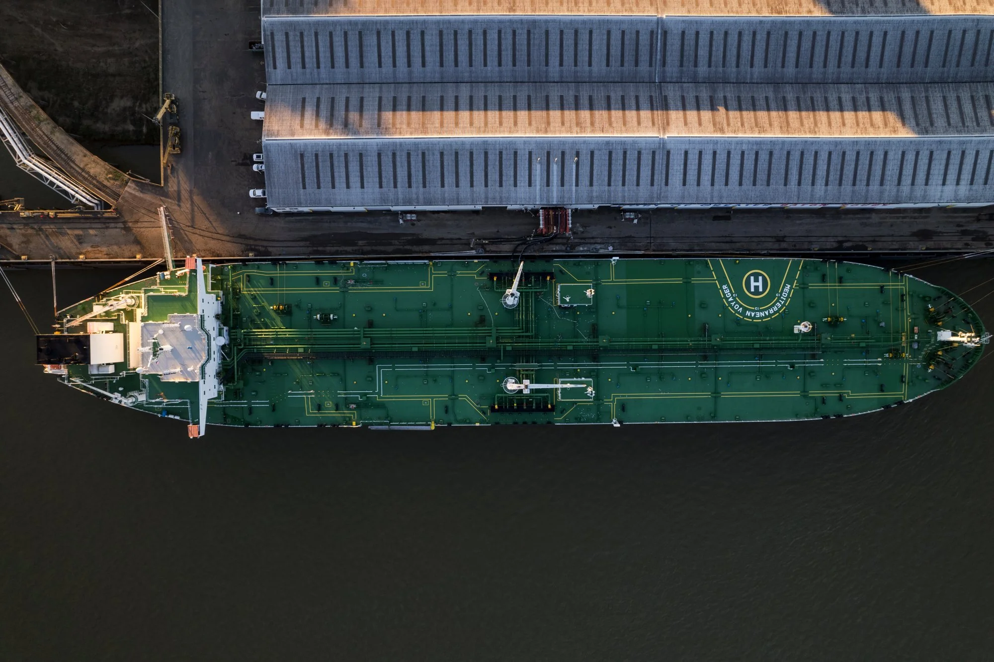 Aerial view of a green cargo ship docked at a harbor with a gray building beside it.