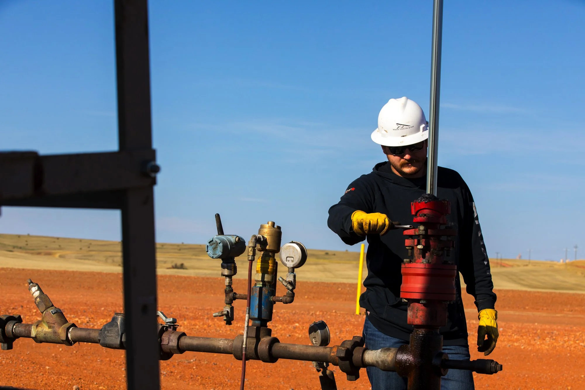 An oil field worker adjusting a valve on a pipeline in a dry, open landscape under a blue sky, wearing safety gear including a white helmet and yellow gloves.