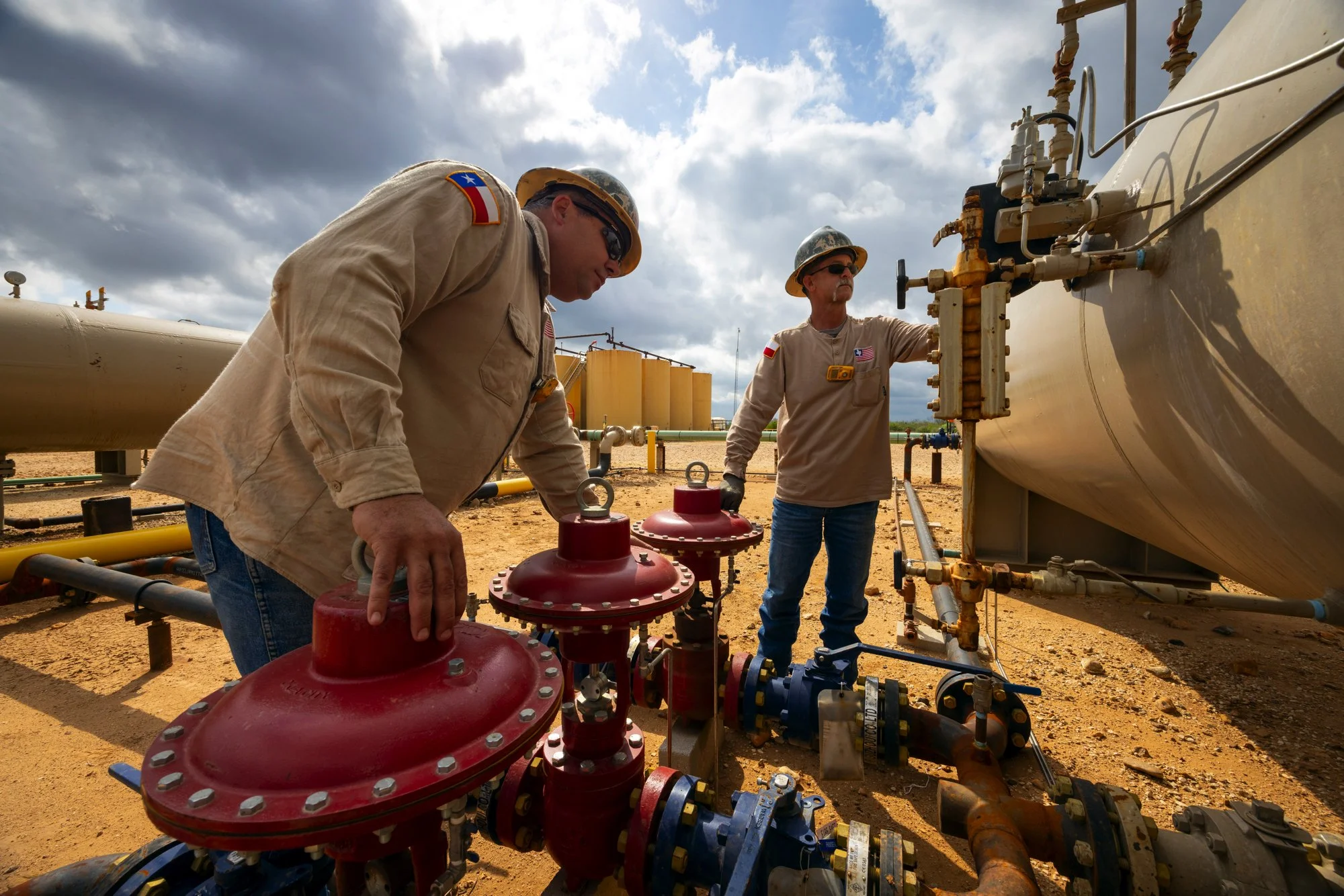 Two workers in tan uniforms and helmets inspecting industrial pipes and valves outdoors on a dirt ground, with a cloudy sky above.