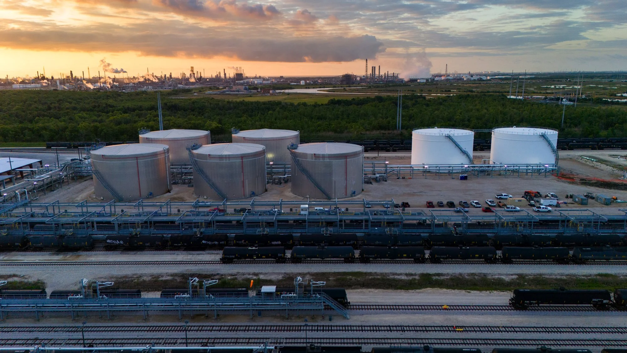 Industrial oil storage tanks and train cars at sunset with smokestacks in the background