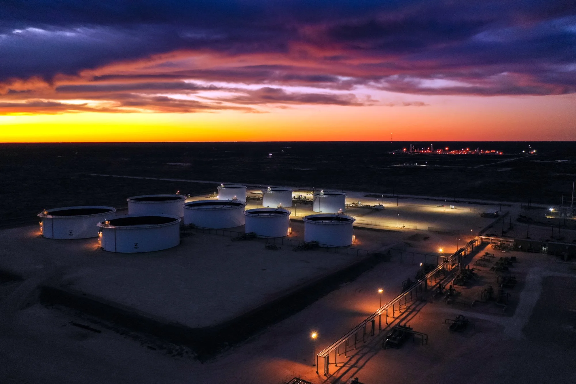 An aerial view of a large industrial fuel storage facility at dusk, with several white cylindrical tanks, illuminated by outdoor lights, and a dark sky with purple and orange clouds.
