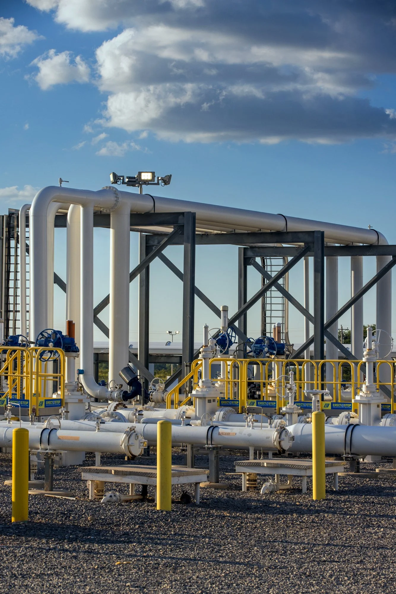 Oil or gas pipeline infrastructure with white pipes, blue valves, yellow safety railings, and a light on top, set outdoors under a blue sky with scattered clouds.