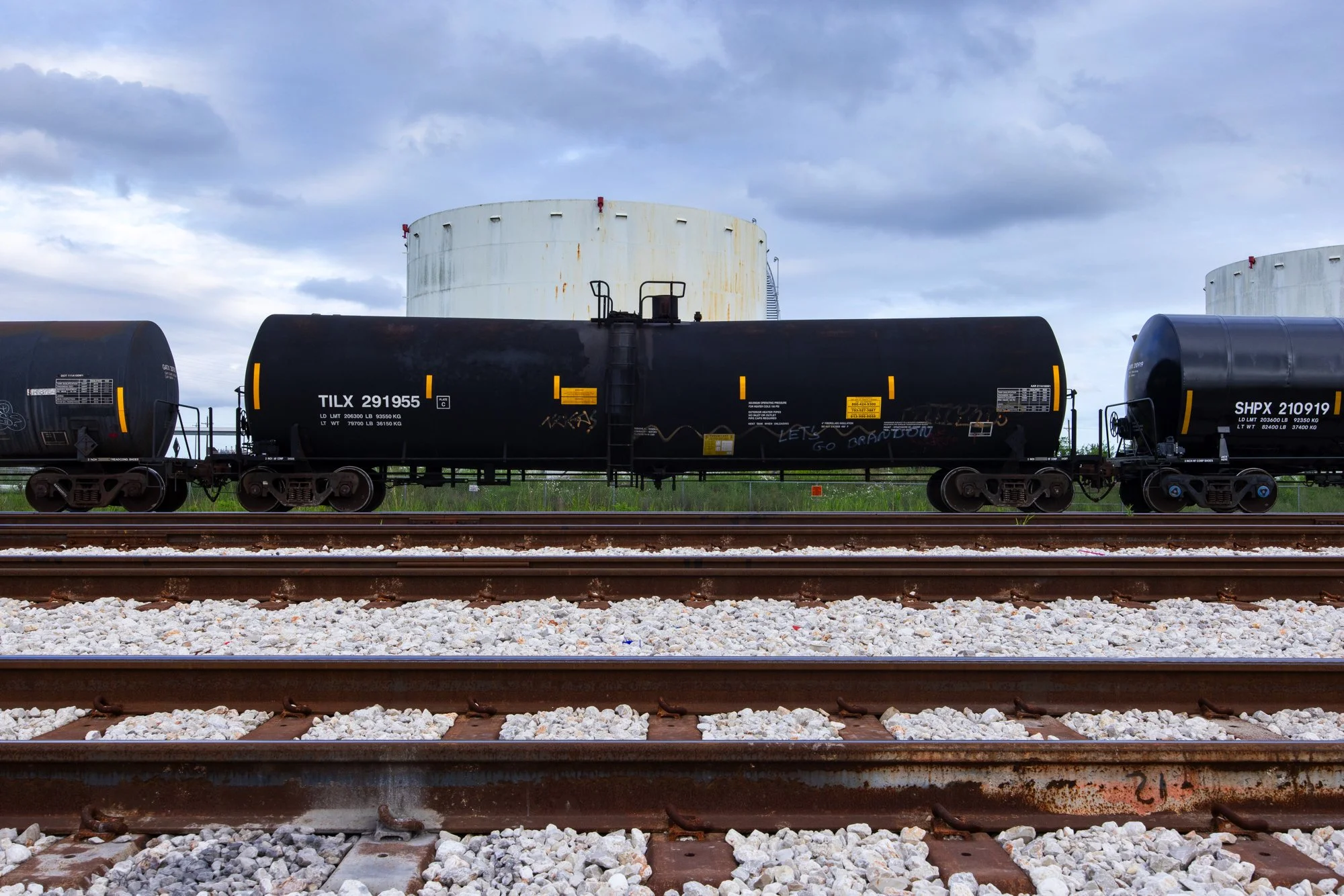 A train with black tank cars on railway tracks under a cloudy sky, with industrial tanks in the background.