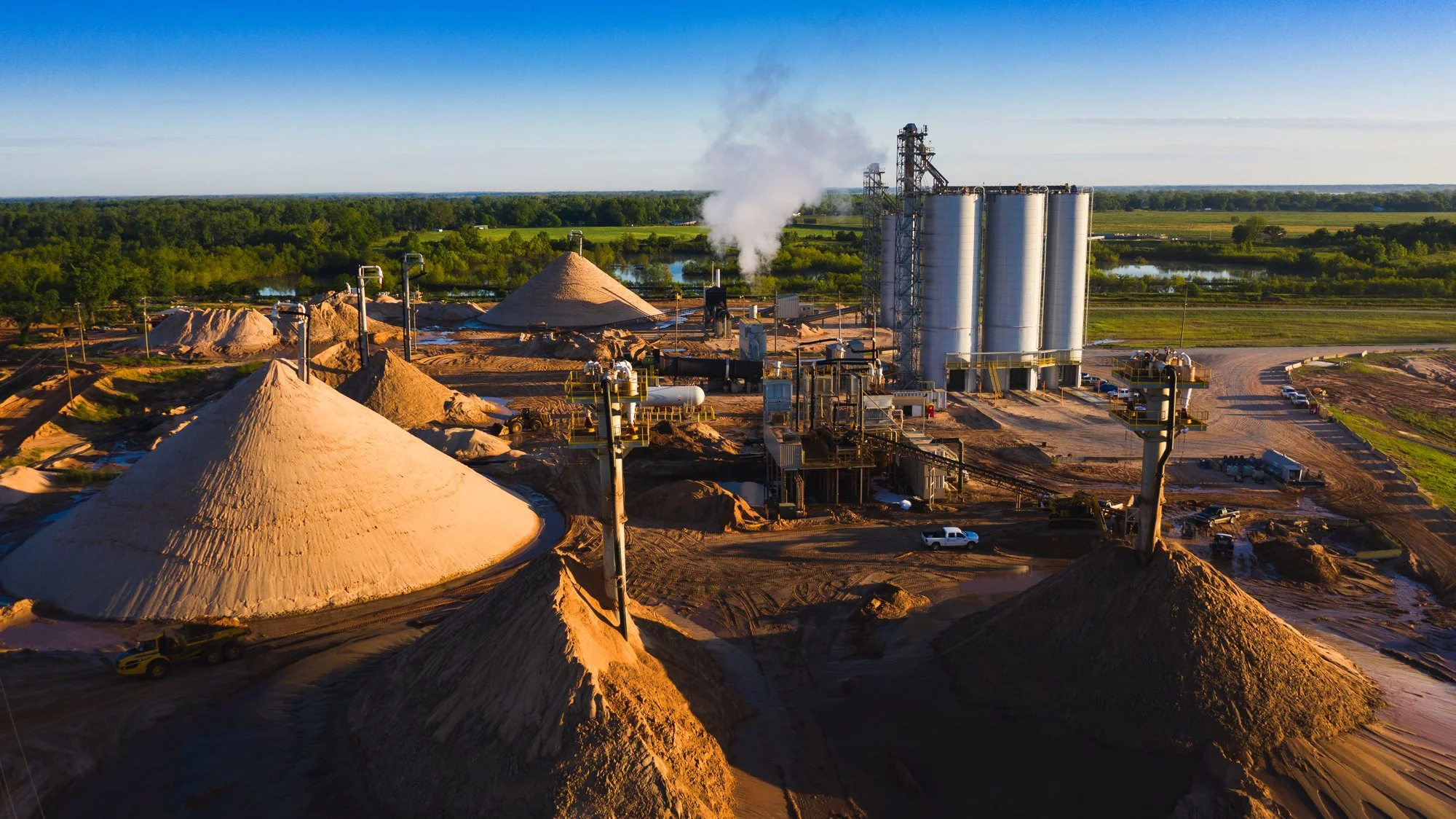 Aerial view of an industrial minerals processing plant with large storage piles, processing equipment, and silos, surrounded by green fields and water bodies.